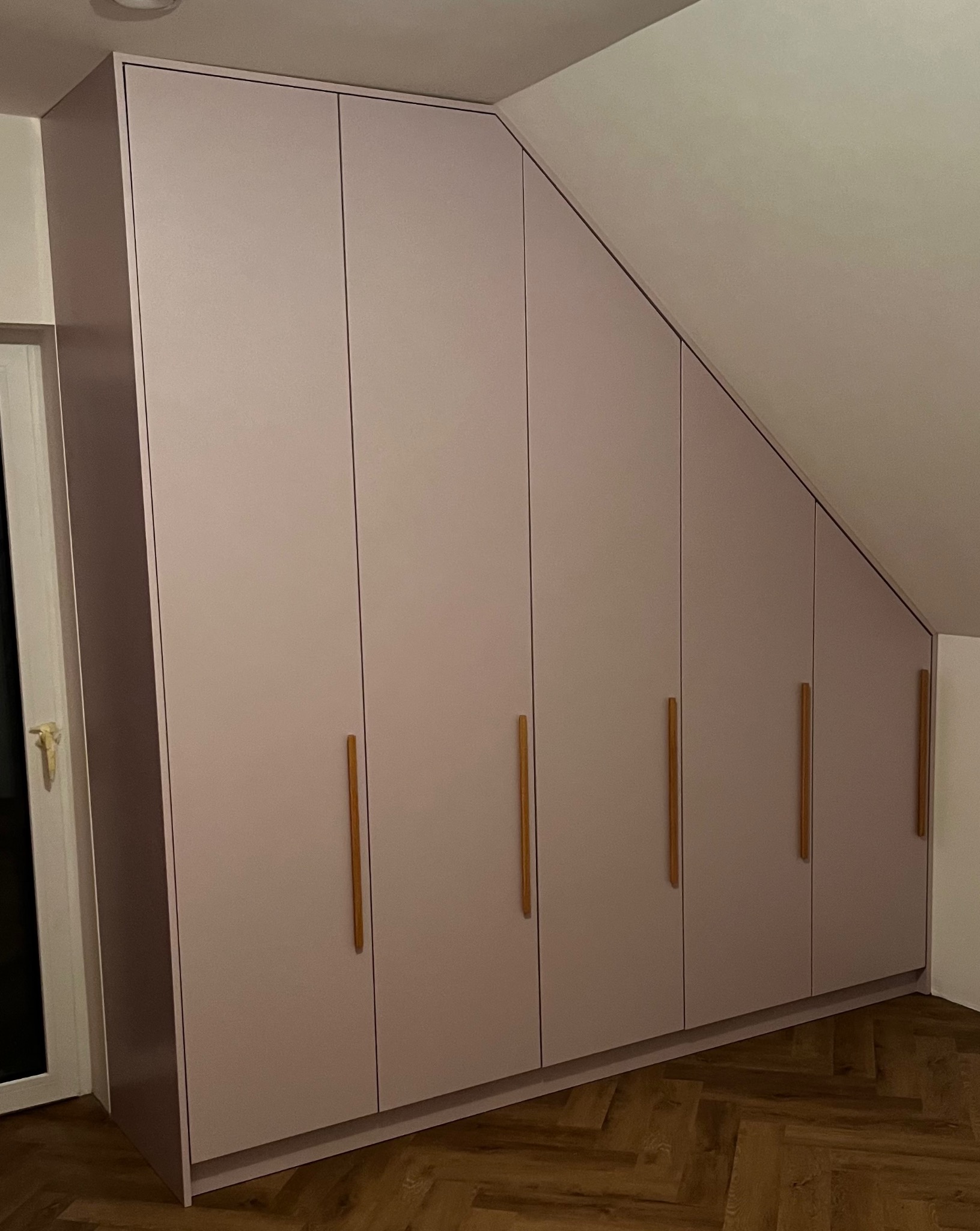 Szafa wnękowa z jasnoróżowymi frontami i drewnianymi uchwytami, dopasowana do skosu dachu na poddaszu.
