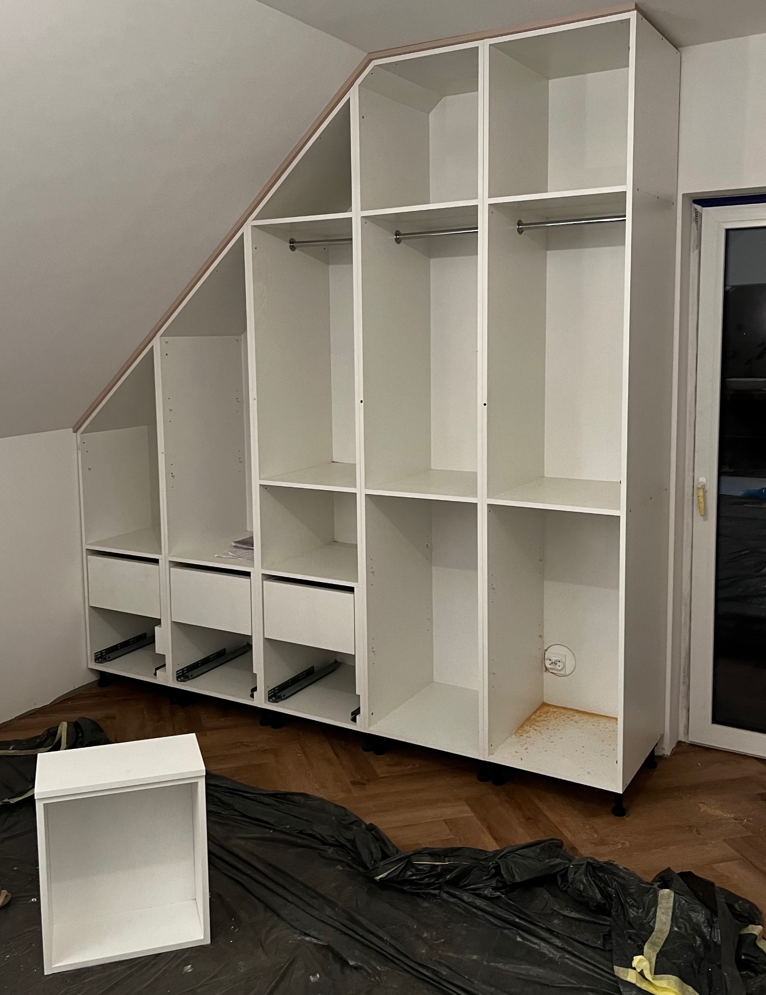 Biała szafa wnękowa z szufladami i półkami, zamontowana na poddaszu ze skosem, w trakcie montażu, widoczne prowadnice szuflad, osłonięta folią podłoga, brakujące drzwiczki szuflady obok.