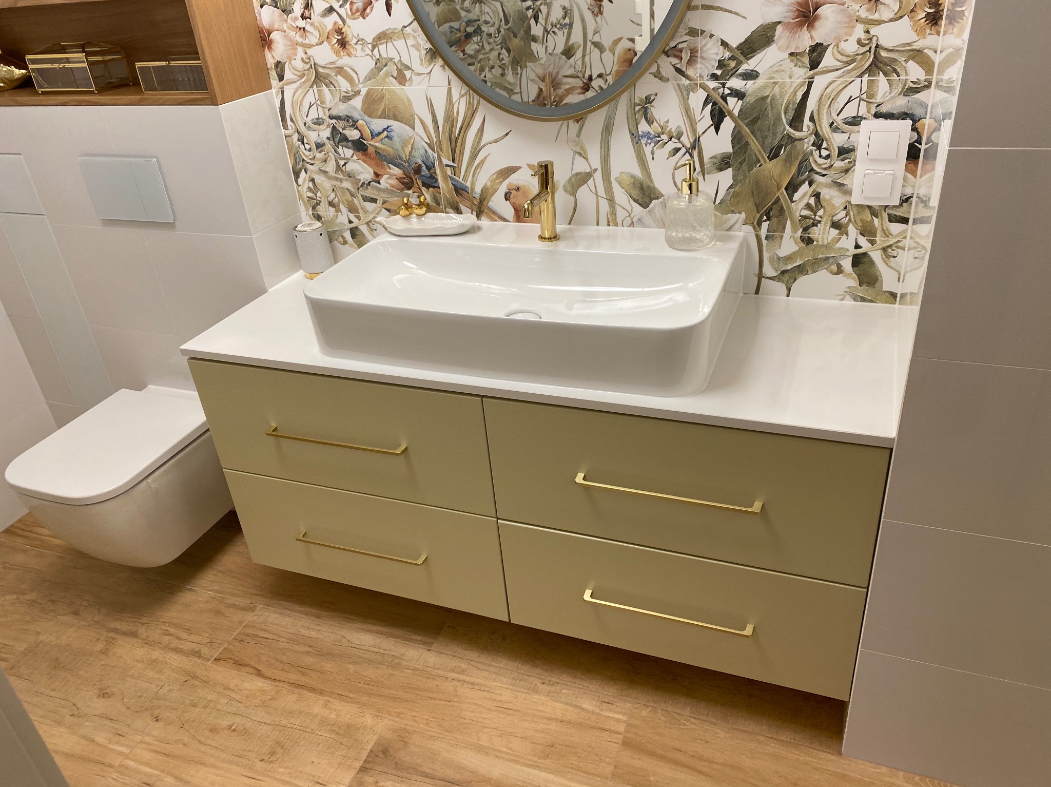 Nowoczesna łazienka z szafką pod umywalką w odcieniach zieleni, białym blatem i umywalką, złote detale armatury i uchwytów, oraz tapeta w motywy roślinne i zwierzęce.