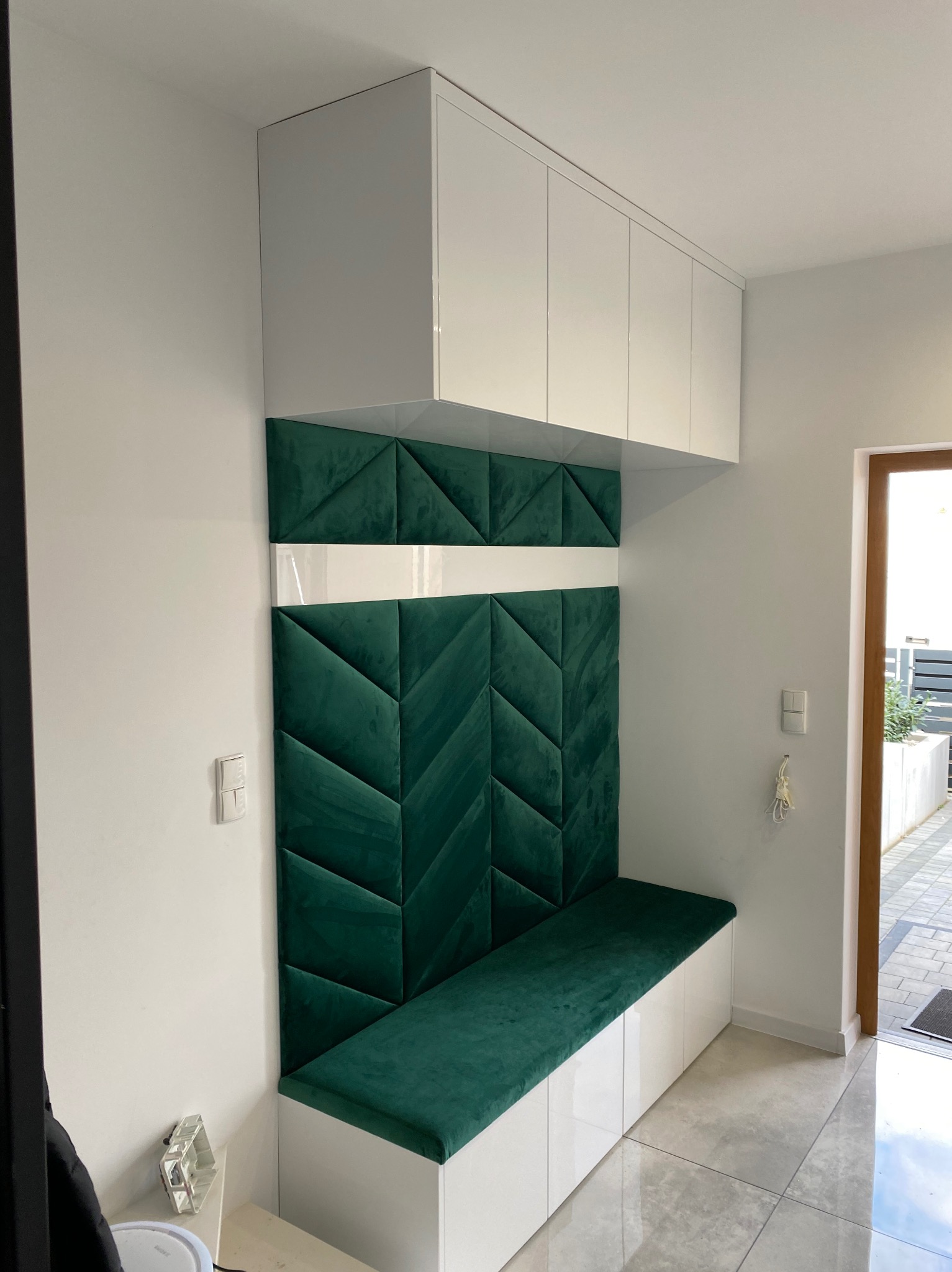 Wnęka z siedziskiem i szafkami w kolorze białym, z tapicerowanym panelem ściennym w kolorze butelkowej zieleni, ułożonym we wzór jodełki. Widoczny fragment podłogi z szarych płytek i drzwi wejściowe.