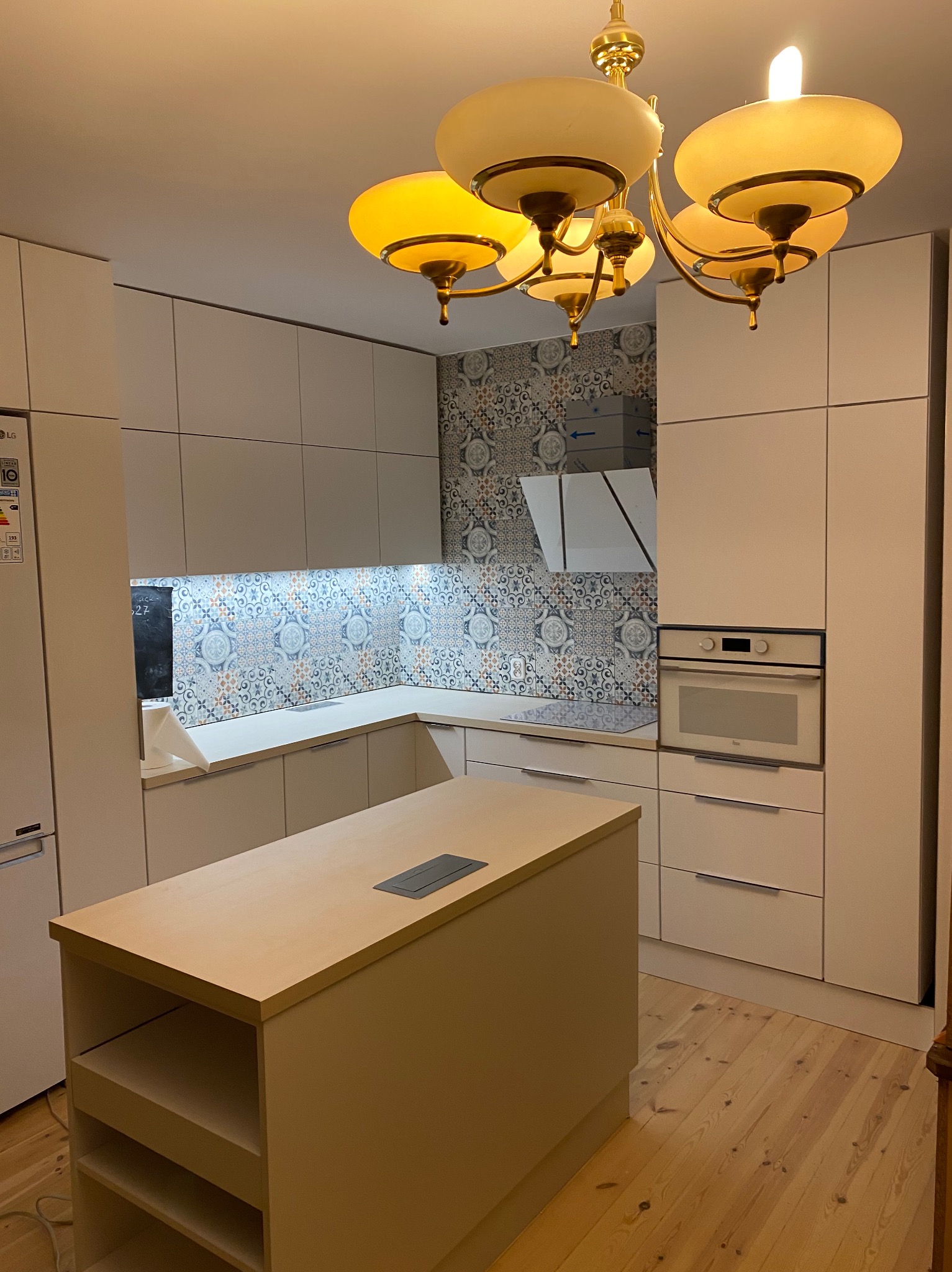 Jasna kuchnia z wyspą, fronty szafek bez uchwytów, kolorowe płytki nad blatem, oświetlenie podszafkowe LED, drewniana podłoga i ozdobny, złoty żyrandol.