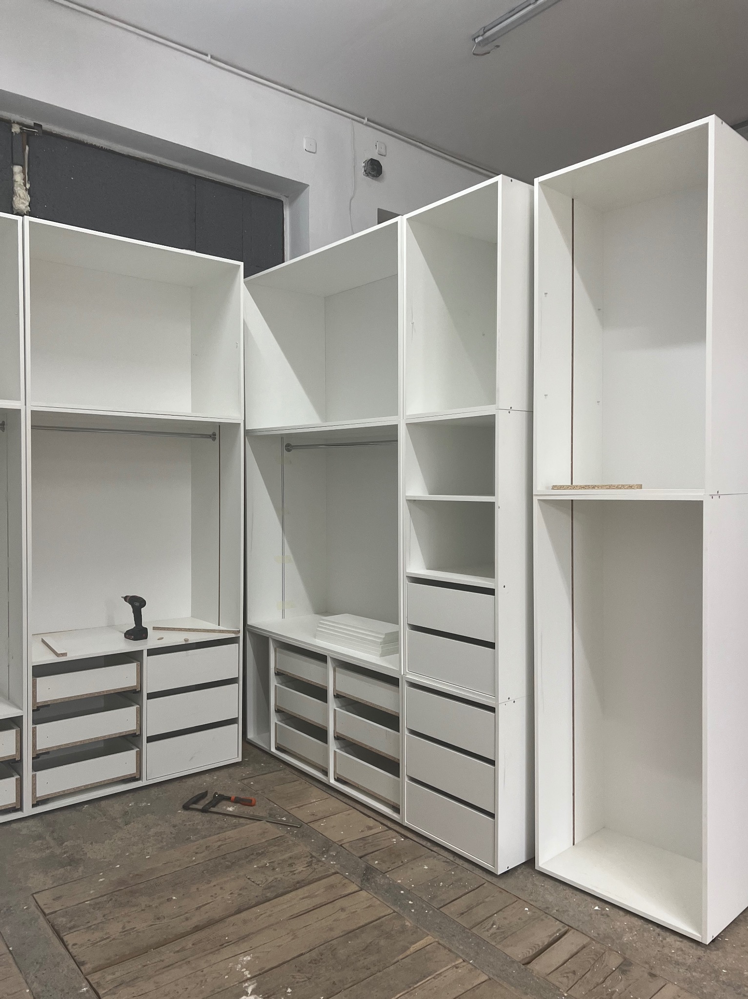 Montaż białych szaf modułowych z szufladami i drążkami na ubrania, widoczny wkrętak akumulatorowy i ścisk stolarski na podłodze z desek podczas prac montażowych.