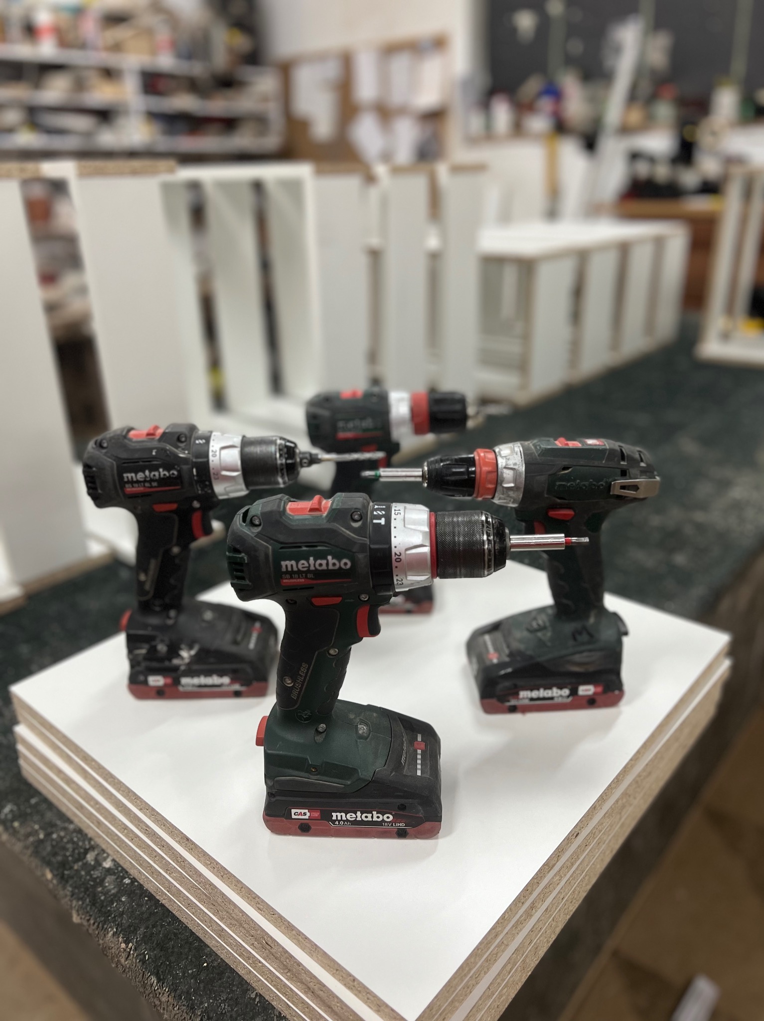 Cztery wiertarko-wkrętarki Metabo ustawione na białych płytach meblowych, warsztat w tle.