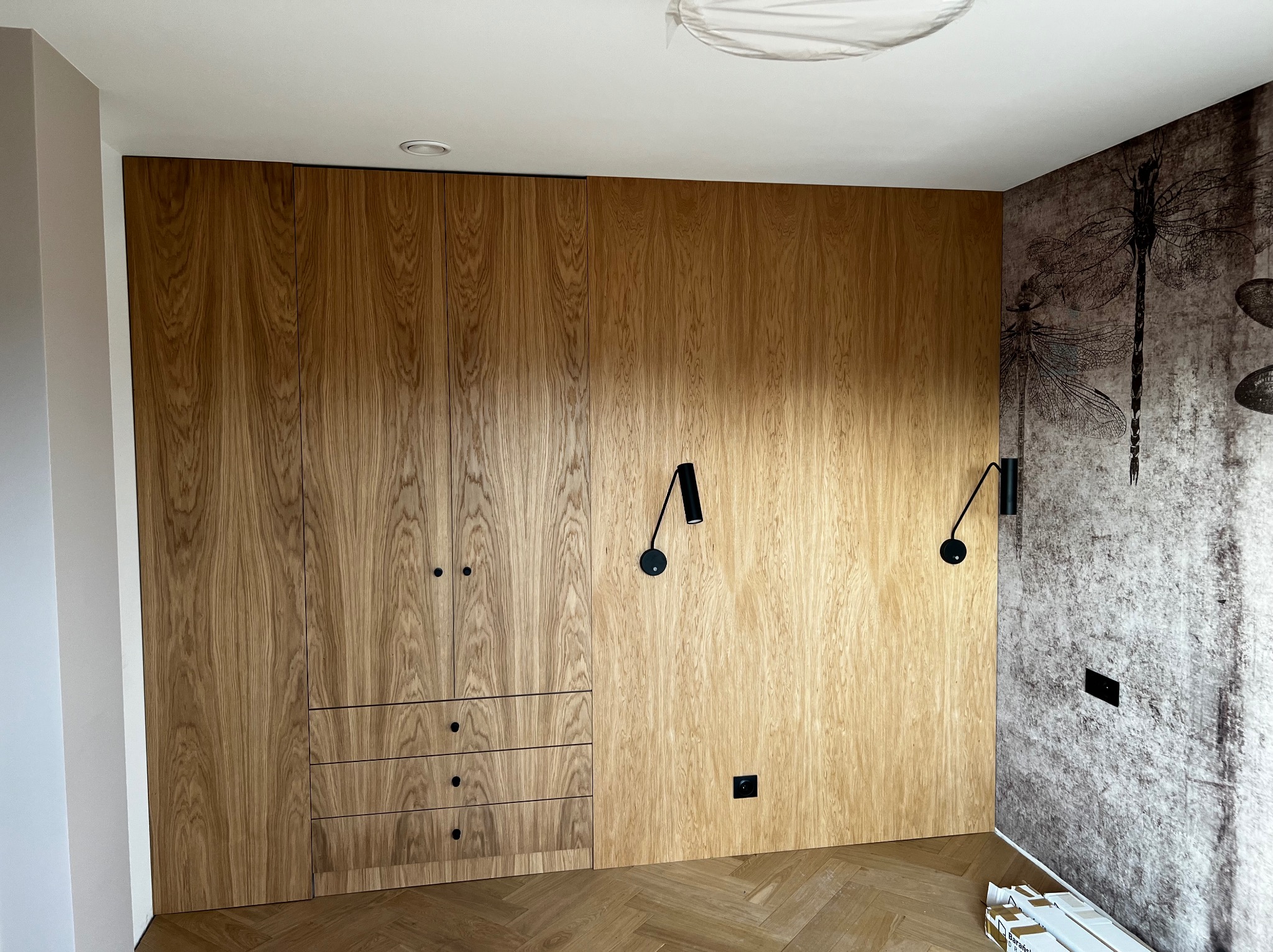 Drewniana zabudowa ściany z szafami, szufladami i ukrytymi drzwiami, oświetlona czarnymi kinkietami, na podłodze parkiet w jodełkę, obok ściana z tapetą w motywy ważek.