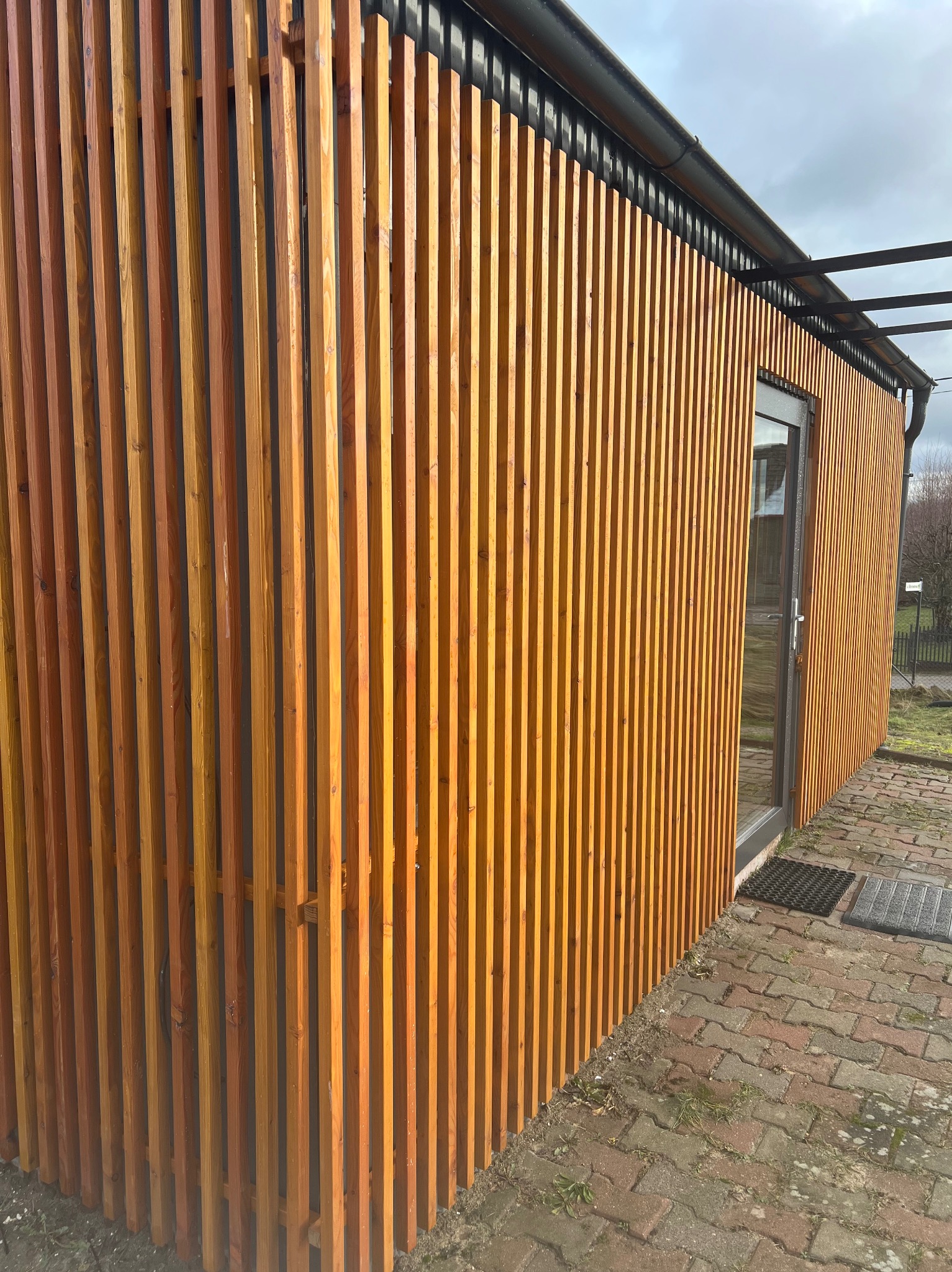 Nowoczesna elewacja budynku z pionowych, drewnianych listew w ciepłym odcieniu brązu, z widocznymi drzwiami wejściowymi i fragmentem chodnika z kostki brukowej.