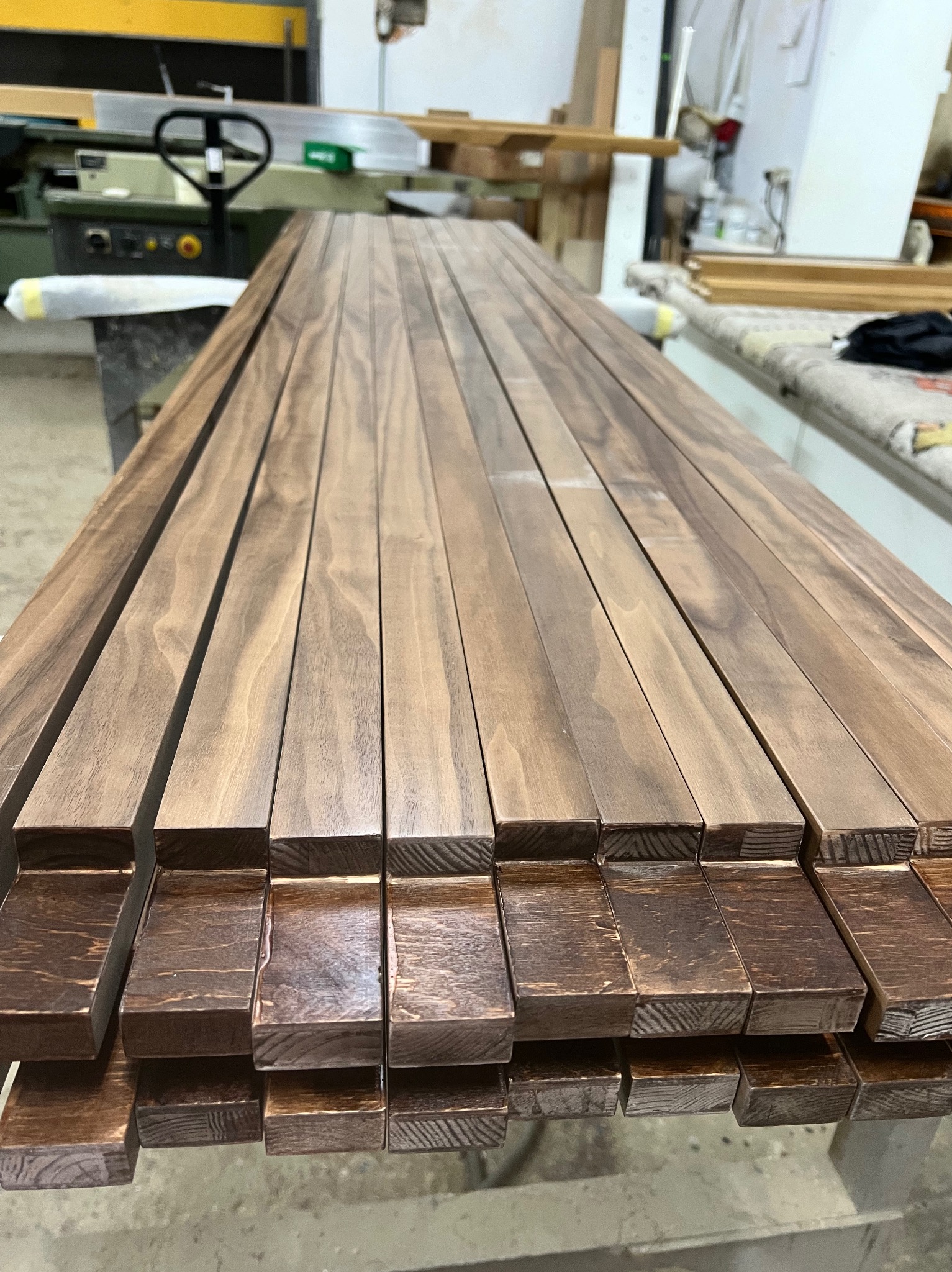 Przygotowane, lakierowane listwy drewniane ułożone w stos, widoczne słoje drewna i maszyny stolarskie w tle.