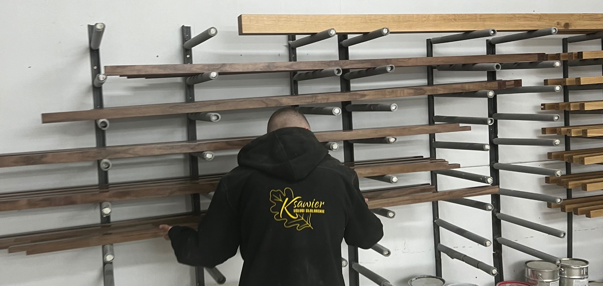 Pracownik w bluzie z logo firmy wybiera z regału drewniane listwy, przygotowane do dalszej obróbki.