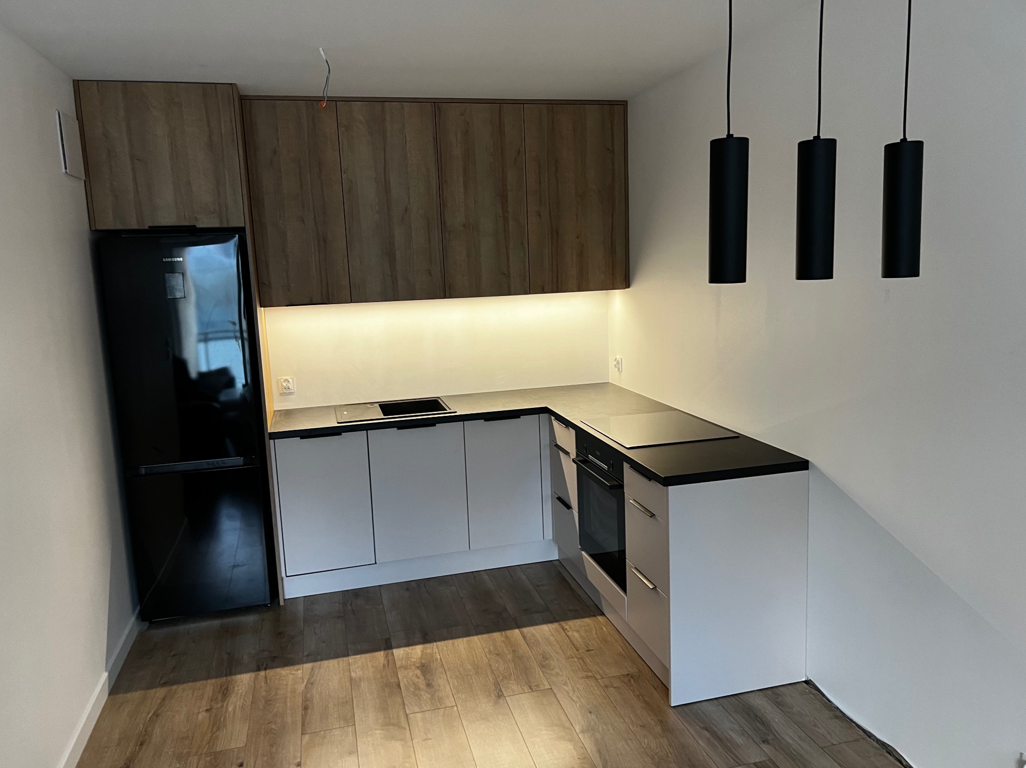 Nowoczesna, narożna zabudowa kuchenna w Luzinie z drewnopodobnymi szafkami górnymi, białymi szafkami dolnymi z czarnym blatem i wiszącymi, cylindrycznymi lampami.