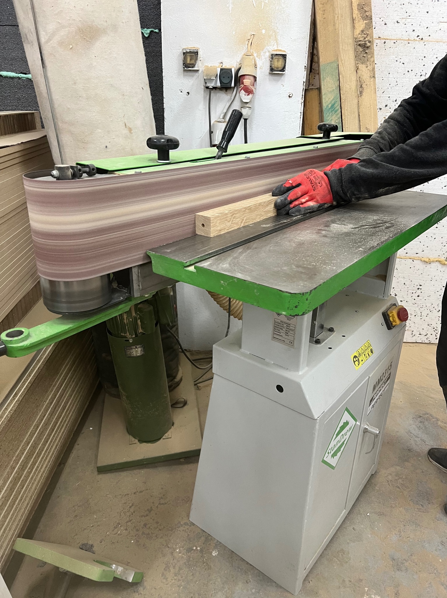 Obróbka drewnianego elementu na szlifierce taśmowej z zielonymi akcentami, operator w czerwonych rękawicach ochronnych, widoczna sterta płyt drewnianych w tle.