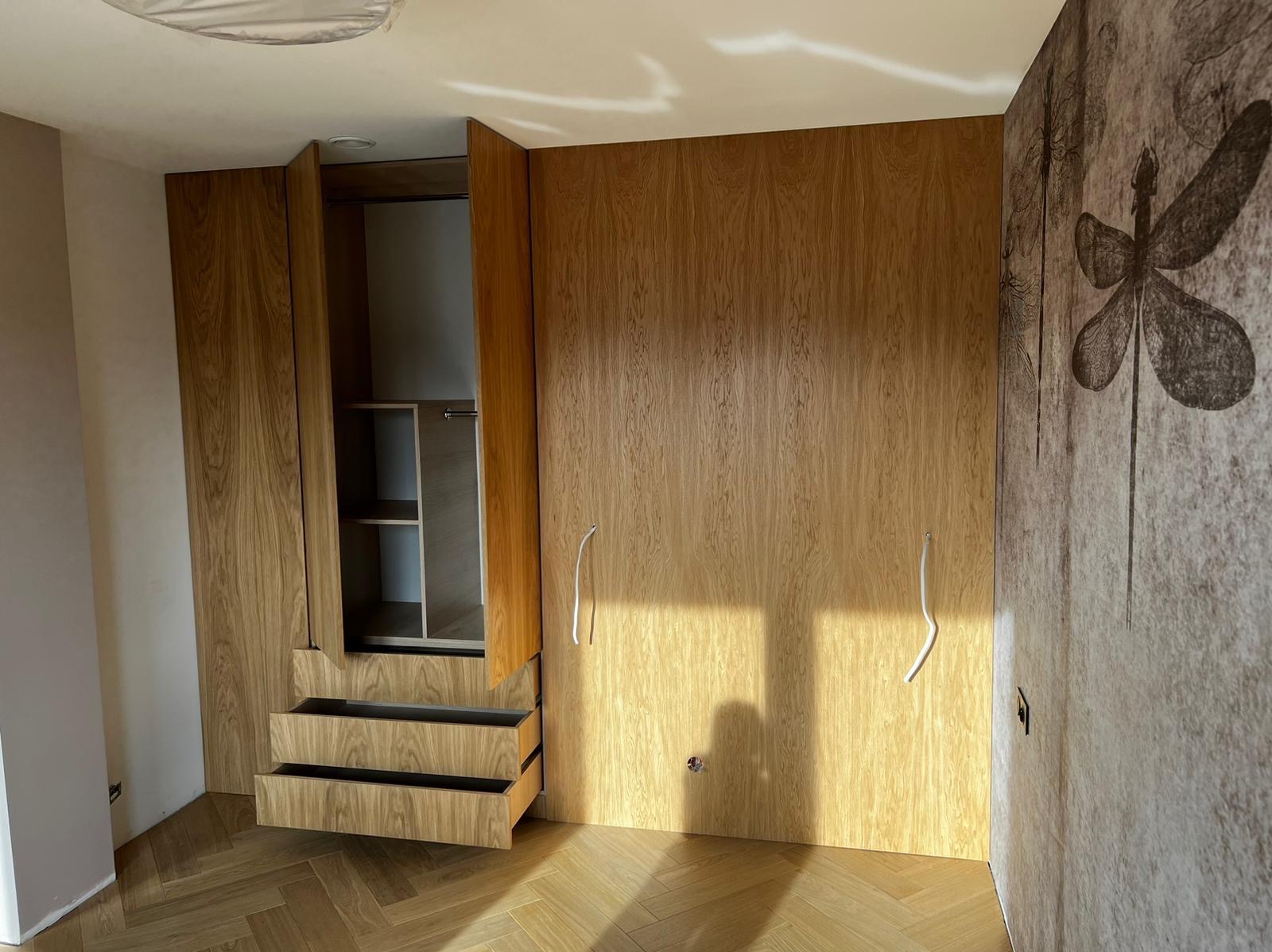 Drewniana szafa wnękowa z otwartymi drzwiami i wysuniętymi szufladami, widoczne półki wewnątrz, na ścianie tapeta z motywem roślinnym, podłoga parkiet w jodełkę, wystające kable elektryczne.