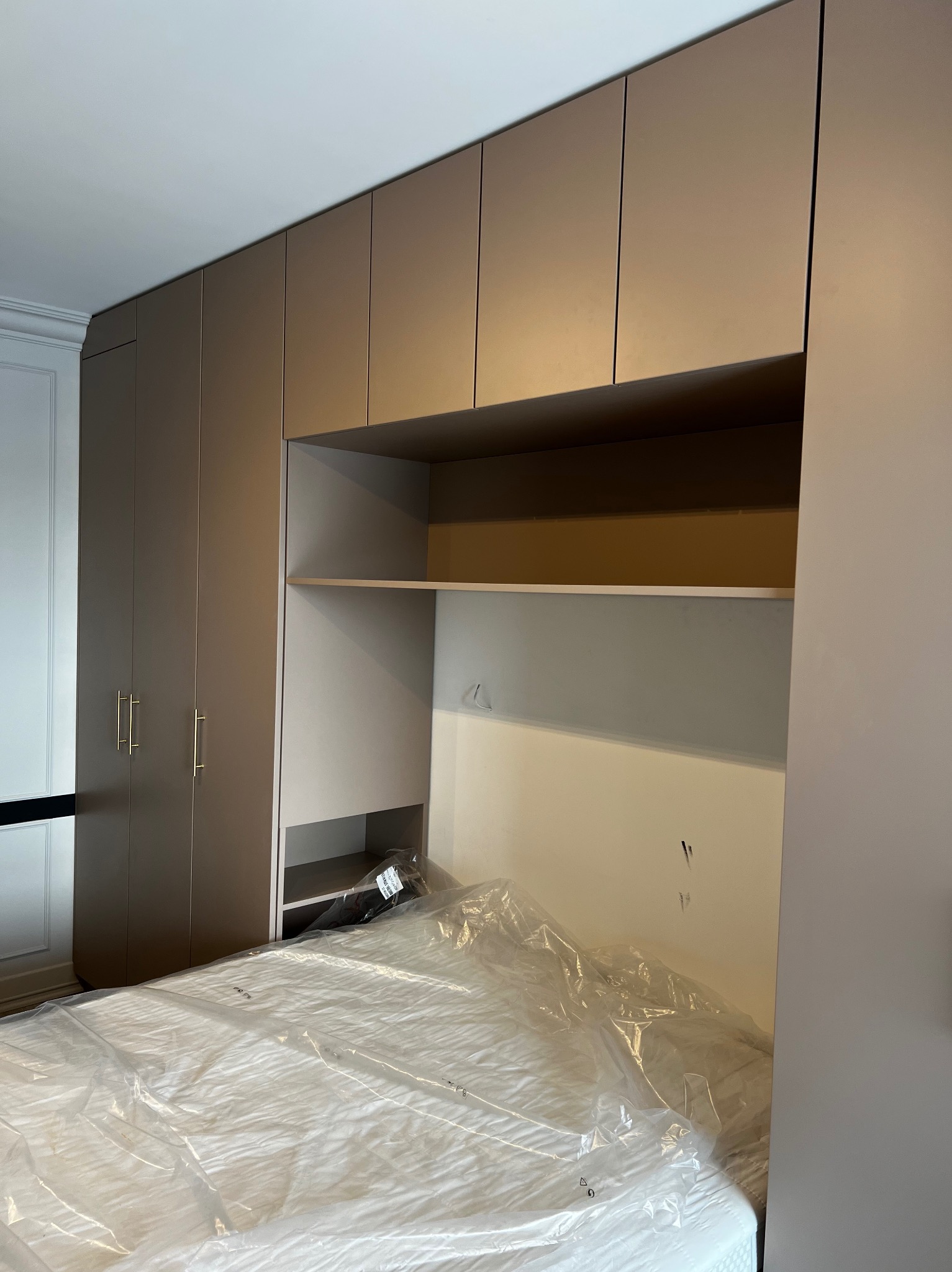 Beżowa zabudowa meblowa z szafami i półkami nad łóżkiem przykrytym folią ochronną, widoczne złote uchwyty szafek i fragment białej sztukaterii.