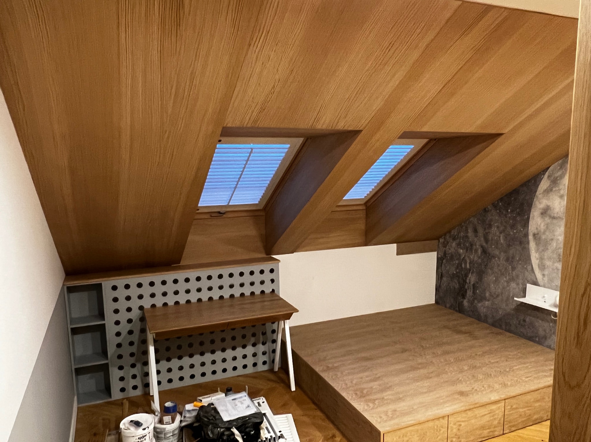 Pokój na poddaszu z drewnianą zabudową obejmującą skośny sufit z oknami dachowymi, biurko z ażurowym panelem, oraz platformę łóżka z szufladami; na podłodze widoczne narzędzia i farby.