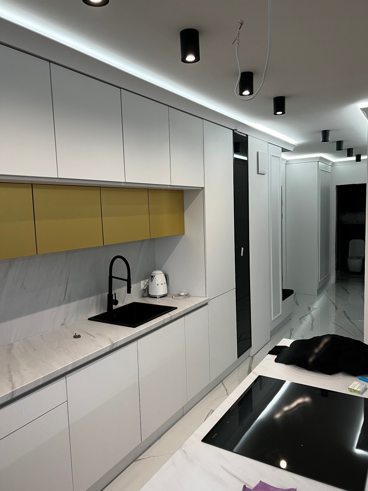 Nowoczesna kuchnia z białymi szafkami, złotymi akcentami i czarną armaturą, widok w głąb korytarza z toaletą.