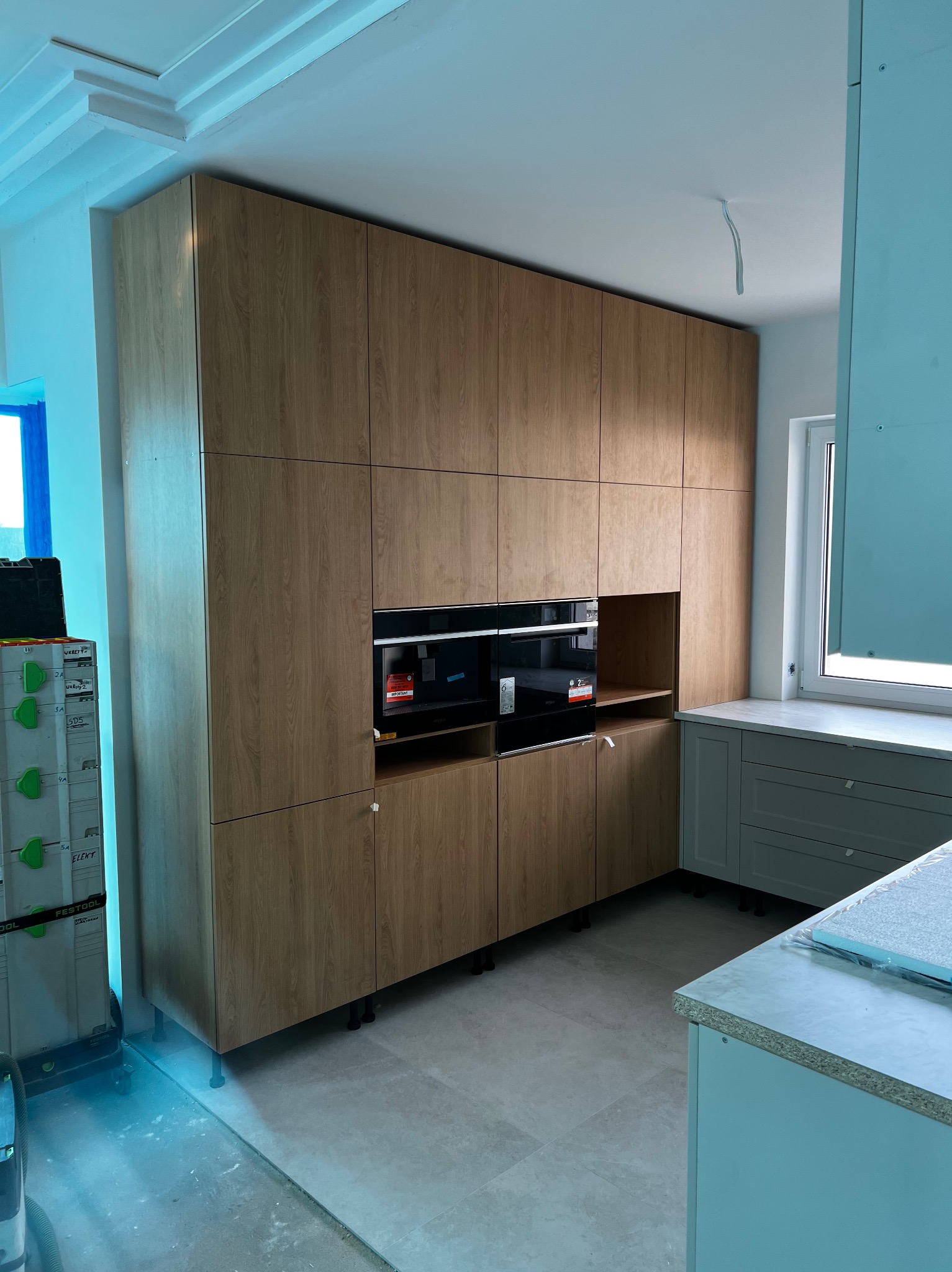 Wysoka zabudowa kuchenna z drewnianymi frontami w odcieniach beżu, z wbudowanymi piekarnikami, w trakcie montażu, widoczne narzędzia i części wyposażenia.