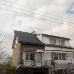 Dom w trakcie ocieplania elewacji styropianem, widoczne rusztowania i balkon z dekoracyjną balustradą z motywem koła wozu.