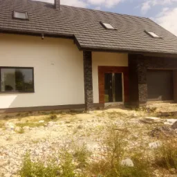 Nowoczesny dom jednorodzinny z białą elewacją i elementami dekoracyjnymi z kamienia, widoczne okna dachowe i brama garażowa, teren wokół domu w trakcie prac wykończeniowych.