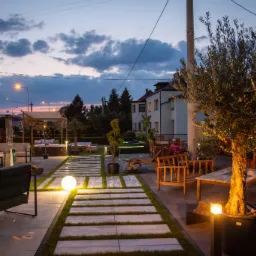 Oświetlony ogród z ścieżką z płyt betonowych i trawy, drewnianymi meblami ogrodowymi, drzewem w donicy i altaną w tle, oświetlenie wieczorne.