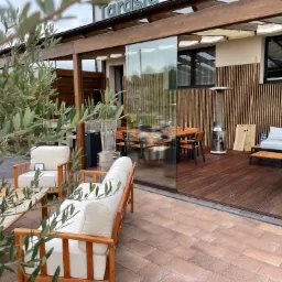 Zadaszony taras z drewnianą pergolą, wyposażony w meble ogrodowe z jasnymi poduchami i stolik z kamiennym blatem, z widokiem na jadalnię na drewnianym podeście i przeszkloną ścianą.