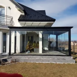 Szklana zabudowa tarasu w Rybniku z czarną ramą, widok z zewnątrz na dom z balkonem i zadbany trawnik.