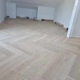 Jasne panele ułożone w jodełkę w przestronnym, jasnym pomieszczeniu z poddaszem. Paczki paneli w folii leżą wzdłuż ścian.