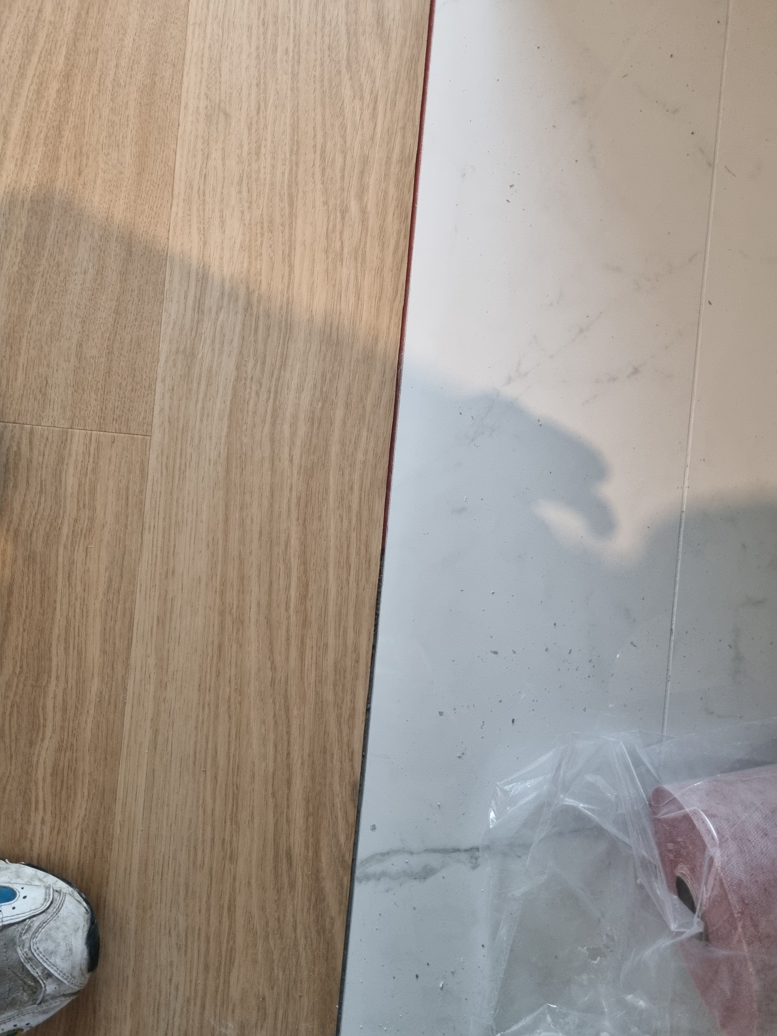 Fragment podłogi z ułożonymi panelami w odcieniach brązu, sąsiadujący z płytkami imitującymi marmur; widoczny but roboczy i rolka podkładu pod panele w folii.