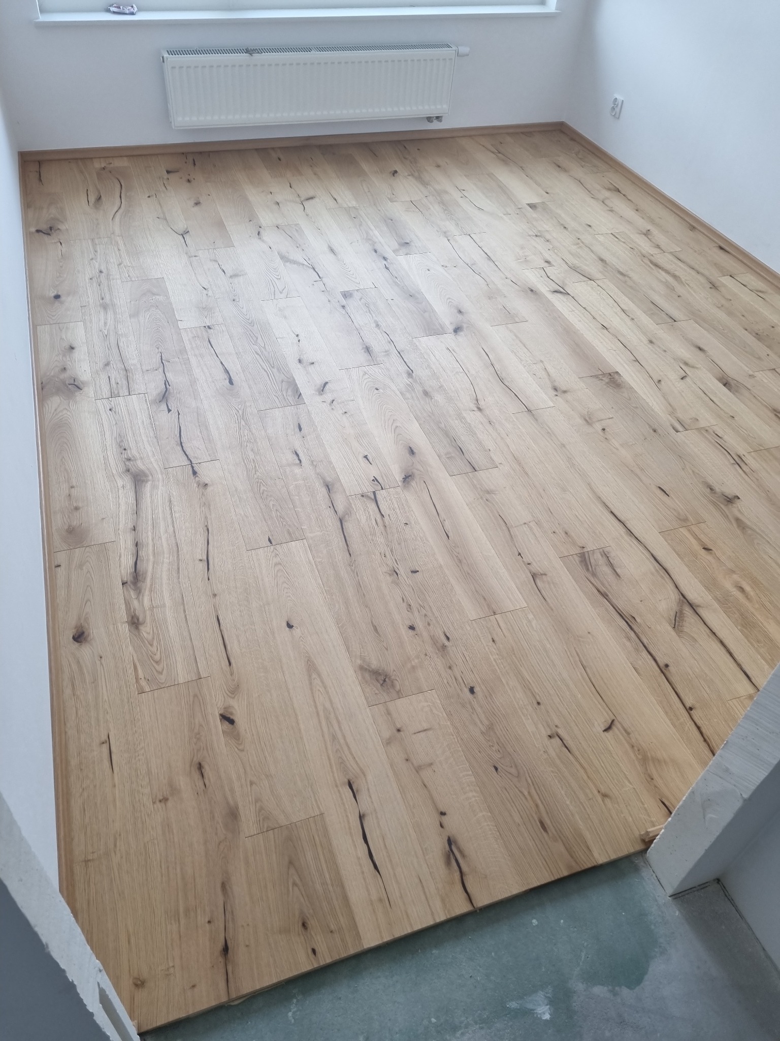 Jasny parkiet z wyraźnym usłojeniem, ułożony w pustym pokoju z białymi ścianami i grzejnikiem, widoczne niedokończone wykończenia przy podłodze.