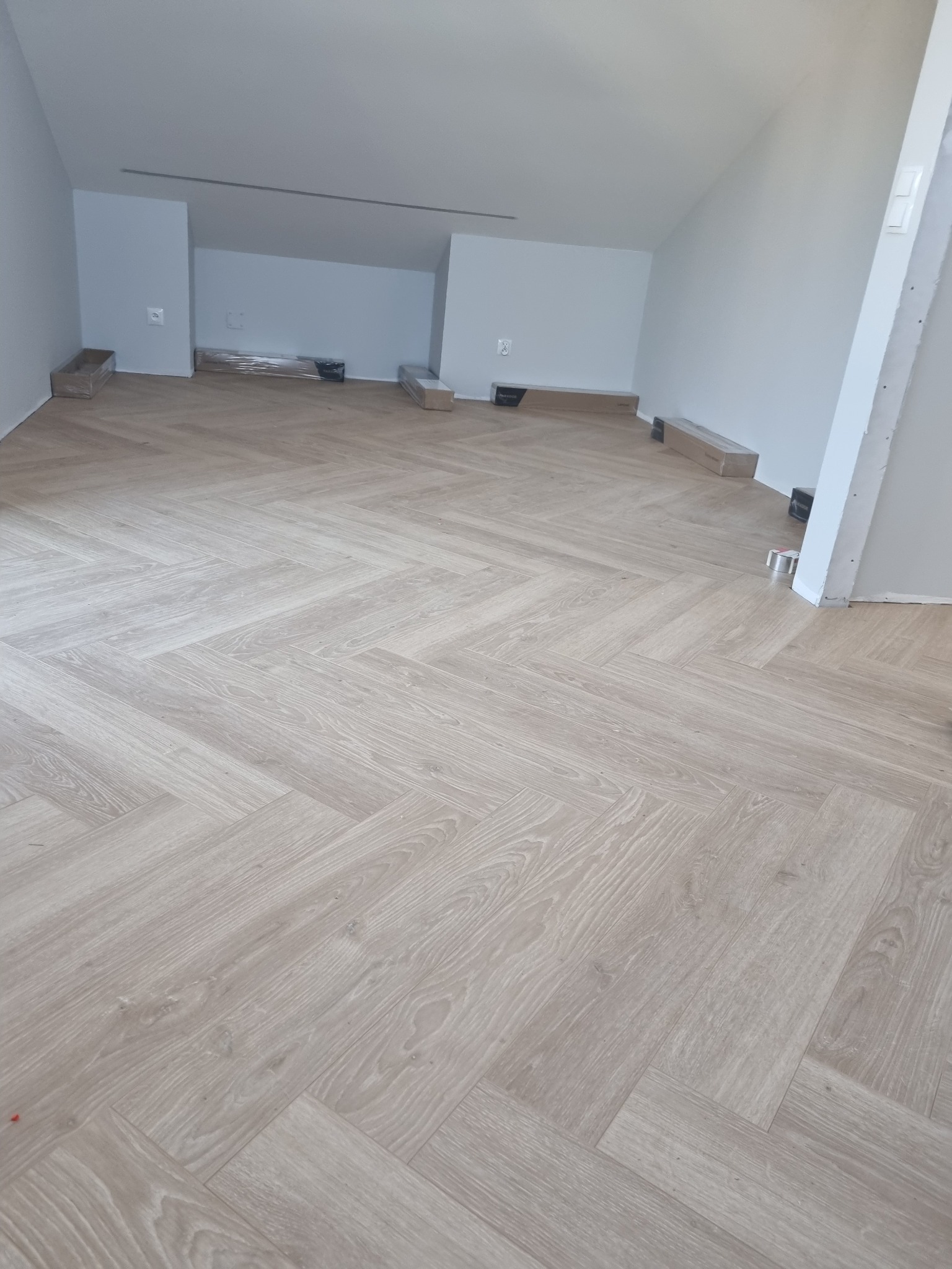 Jasne panele ułożone w jodełkę w przestronnym, jasnym pomieszczeniu z poddaszem. Paczki paneli w folii leżą wzdłuż ścian.