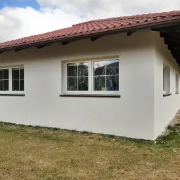 Świeżo otynkowany, biały dom parterowy z brązową dachówką, widok z boku na trawnik z opadłymi puchem topoli.