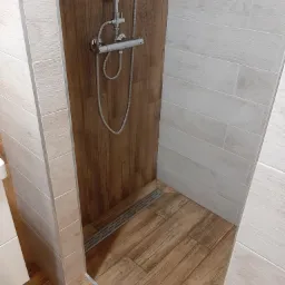 Nowoczesna kabina prysznicowa z odpływem liniowym, wykończona płytkami imitującymi drewno w dwóch odcieniach, jasne na ścianach i ciemniejsze na podłodze oraz tylnej ścianie kabiny.