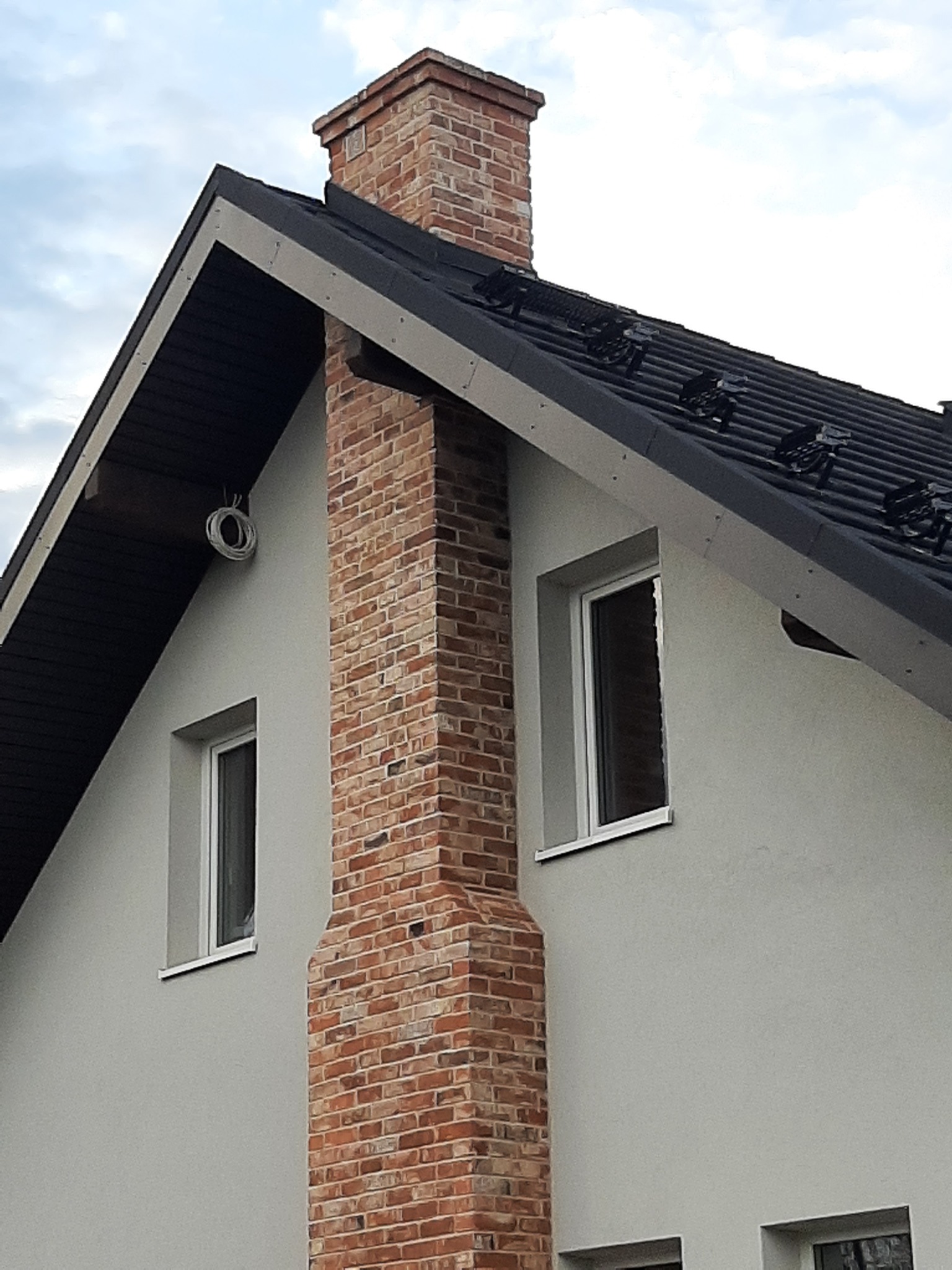Fragment elewacji domu z ceglanym kominem, widoczne okna z białymi ramami i zwój kabla na ścianie.