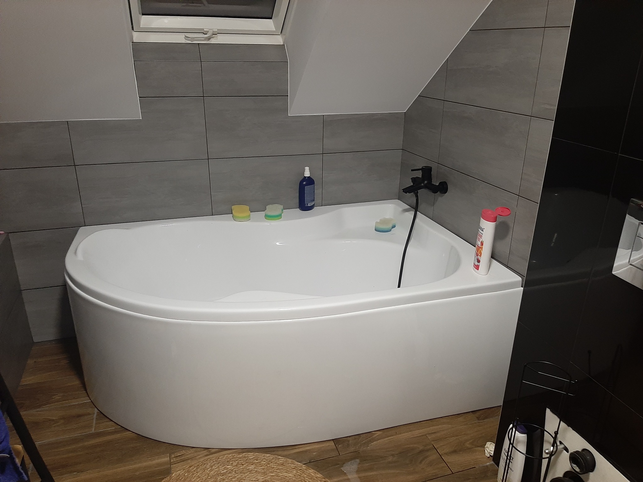 Nowoczesna łazienka z białą, zaokrągloną wanną, szarymi płytkami na ścianach i drewnopodobną podłogą, z czarną armaturą i oknem dachowym.