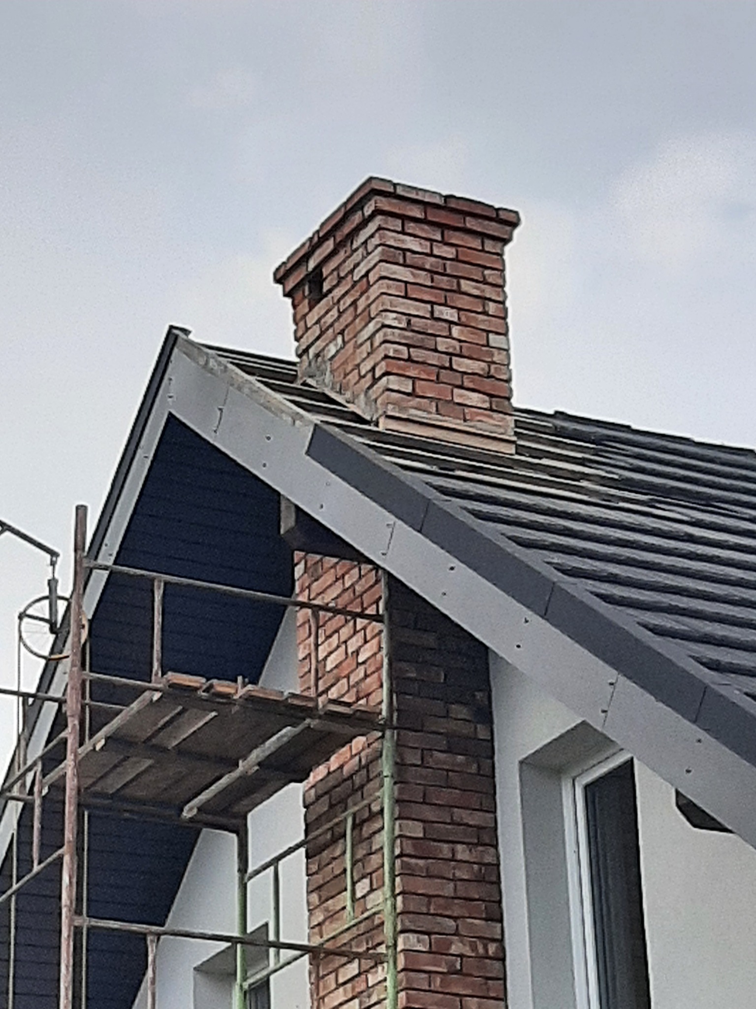 Murowany komin z cegły, fragment dachu z szarą dachówką i białą elewacją, widoczne rusztowanie.