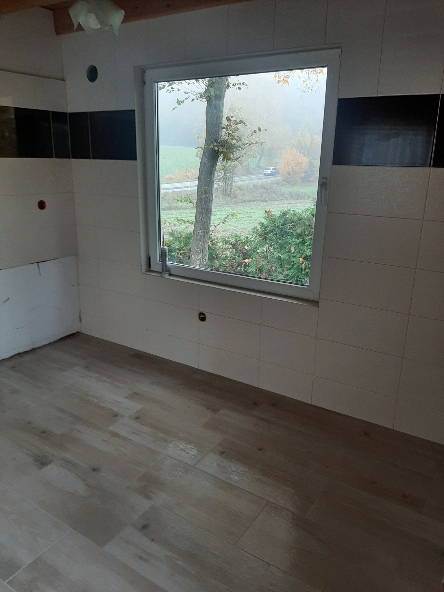 Wnętrze łazienki w trakcie remontu, z ułożonymi płytkami podłogowymi imitującymi drewno i częściowo wykończonymi ścianami z białych płytek z czarnym pasem dekoracyjnym, widok na zewnątrz przez okno.