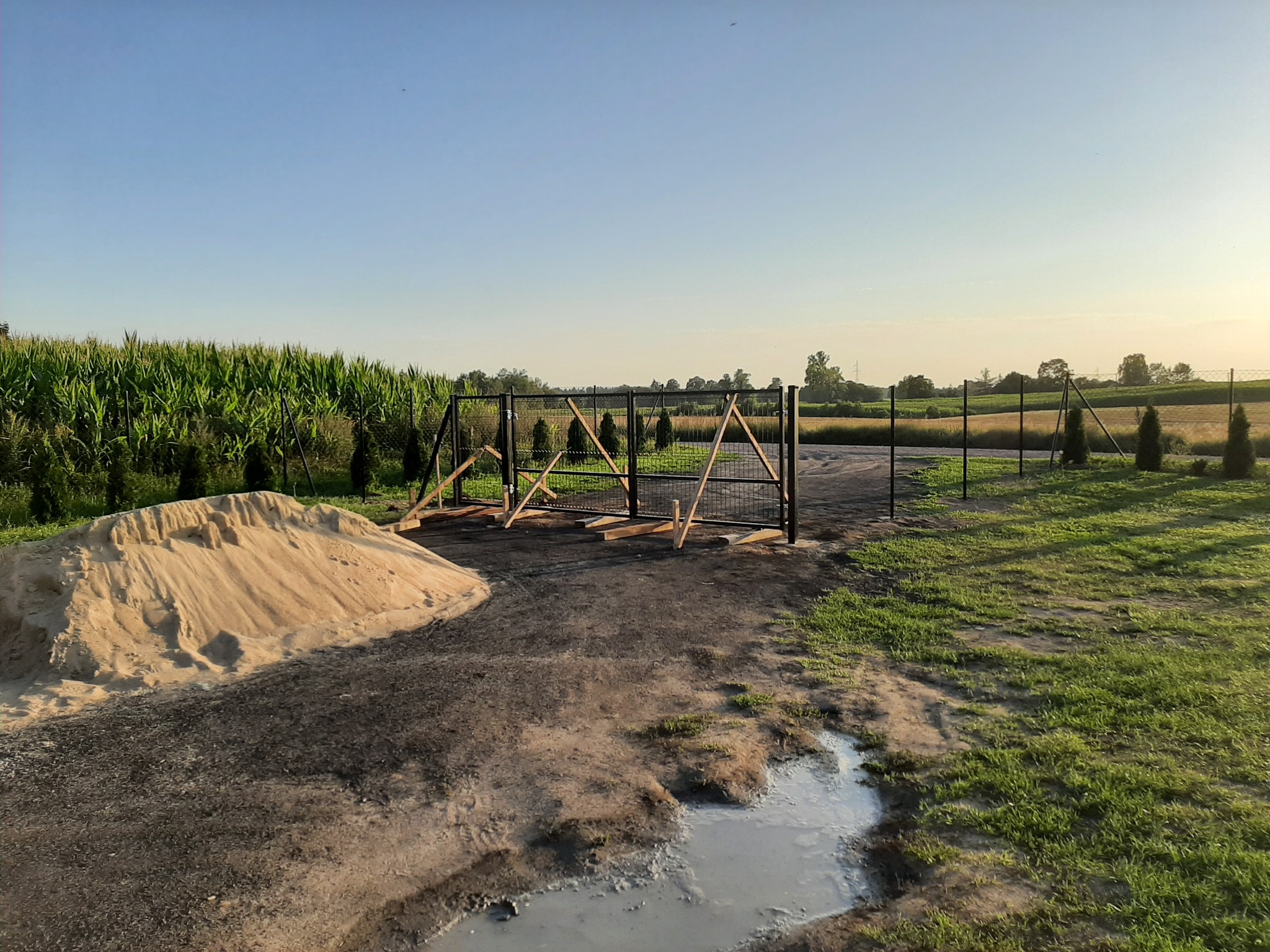Montaż metalowej bramy wjazdowej na posesję, widoczna sterta piasku i tymczasowe drewniane podpory, w tle pole kukurydzy i drzewa.