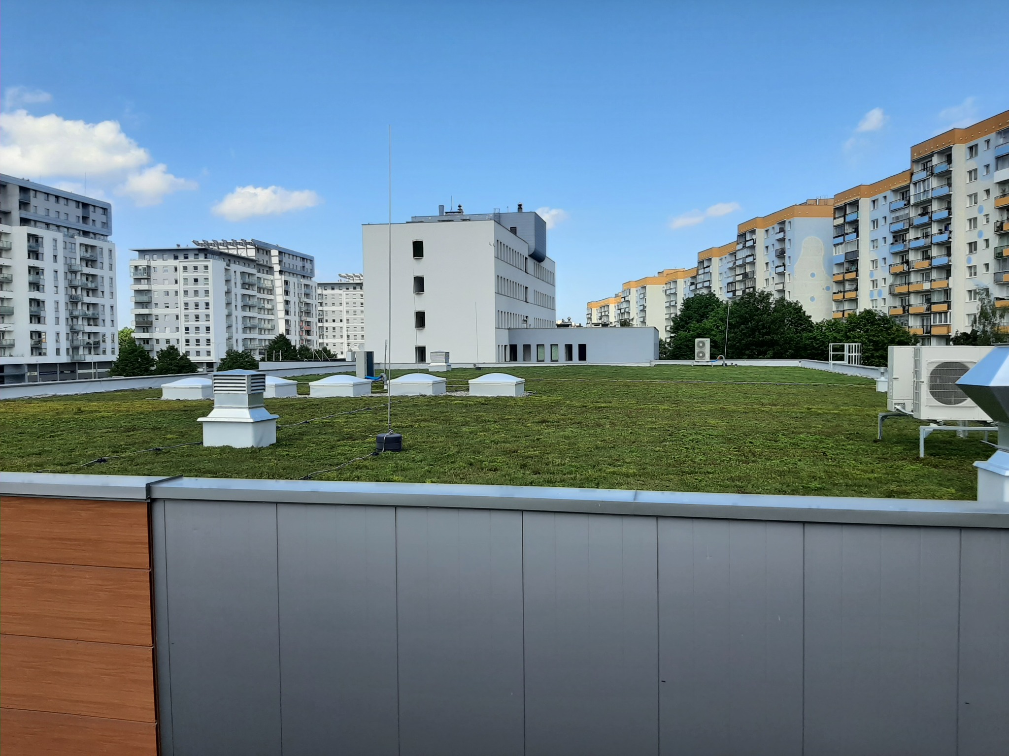 Widok z góry na budynki mieszkalne i biurowe z zielonym dachem, widoczne kominy wentylacyjne, klimatyzatory i anteny, błękitne niebo z chmurami.