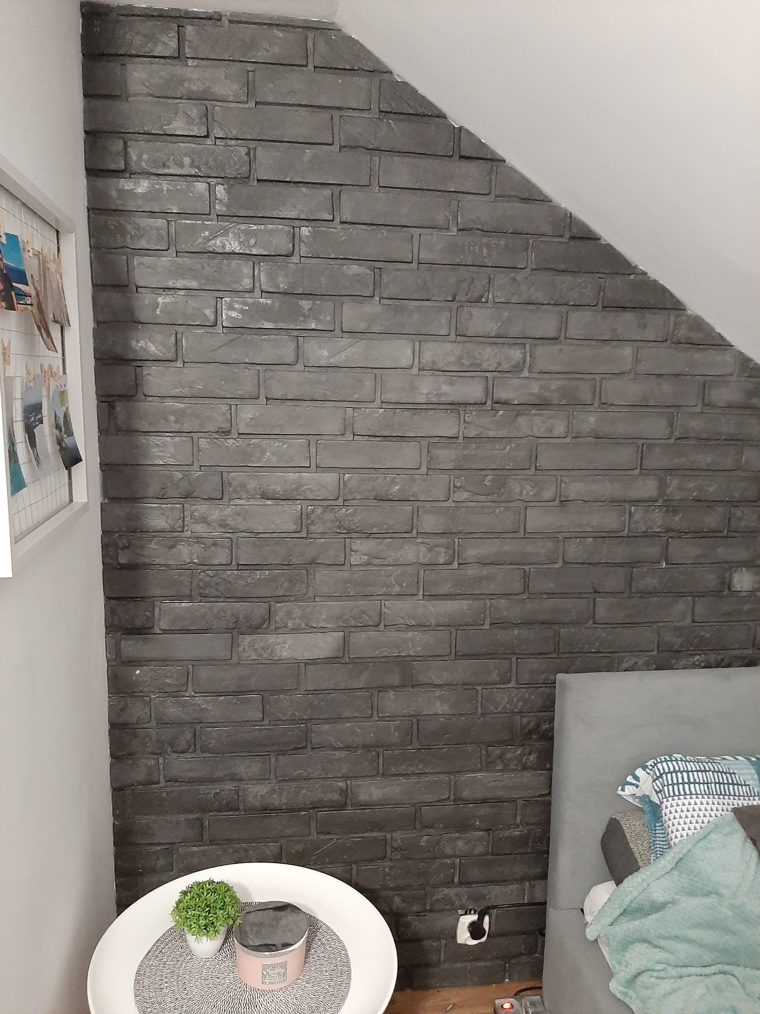 Ściana wykończona szarą cegłą dekoracyjną w pokoju na poddaszu, widoczny stolik z rośliną i świecą oraz fragment łóżka.