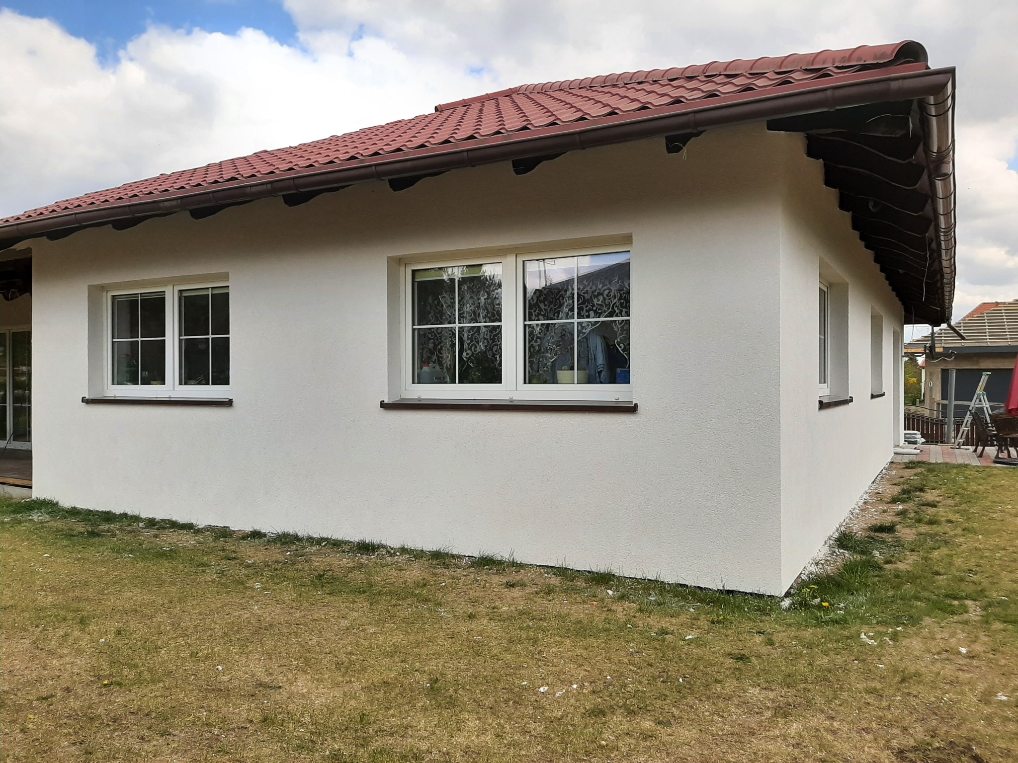 Świeżo otynkowany, biały dom parterowy z brązową dachówką, widok z boku na trawnik z opadłymi puchem topoli.