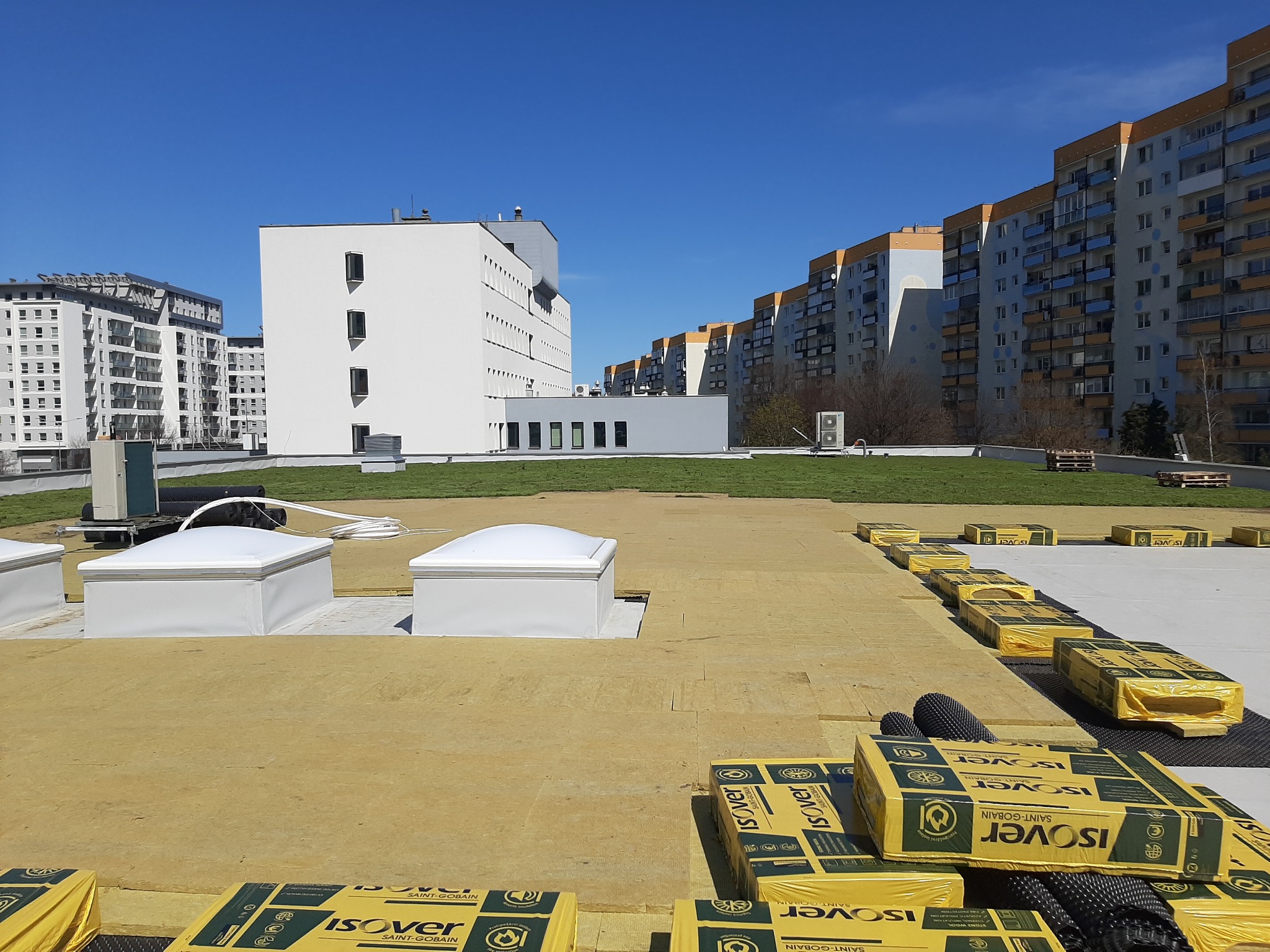 Izolacja dachu płaskiego wełną mineralną Isover, widoczne świetliki dachowe i budynki mieszkalne w tle.