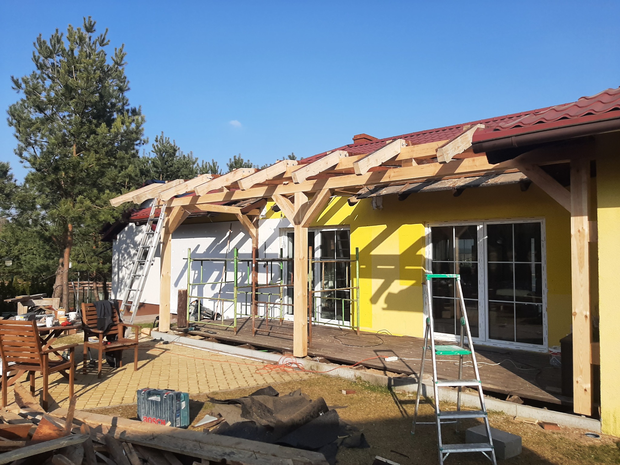 Budowa drewnianej pergoli przy żółtej elewacji domu z czerwonym dachem, widoczne rusztowania, drabiny i narzędzia, w tle drzewa i błękitne niebo.
