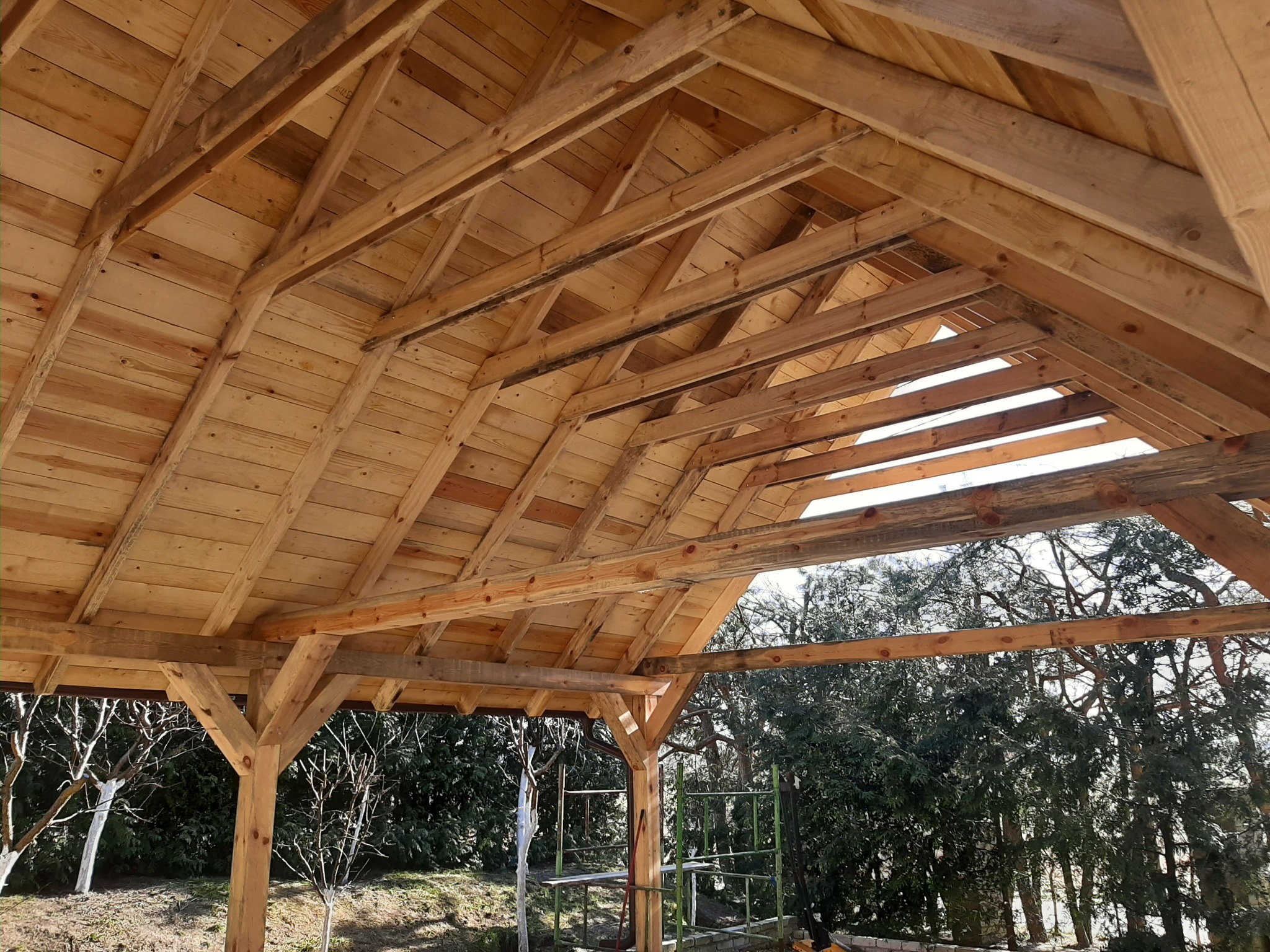 Drewniana konstrukcja zadaszenia w surowym stanie, widok od dołu, z widocznymi elementami więźby dachowej i otaczającą zielenią w tle.