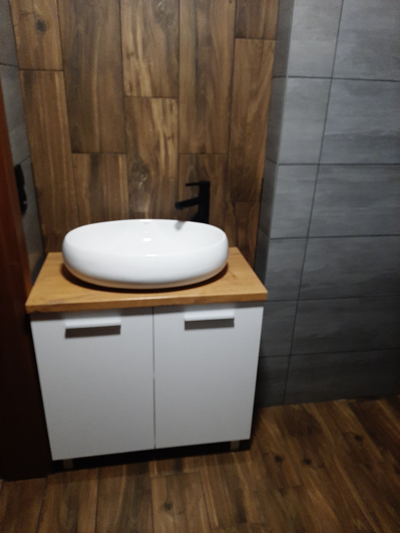 Nowoczesna łazienka z okrągłą, białą umywalką nablatową na szafce z drewnianym blatem, czarna bateria, drewnopodobne płytki na ścianie i podłodze oraz szare płytki imitujące beton.