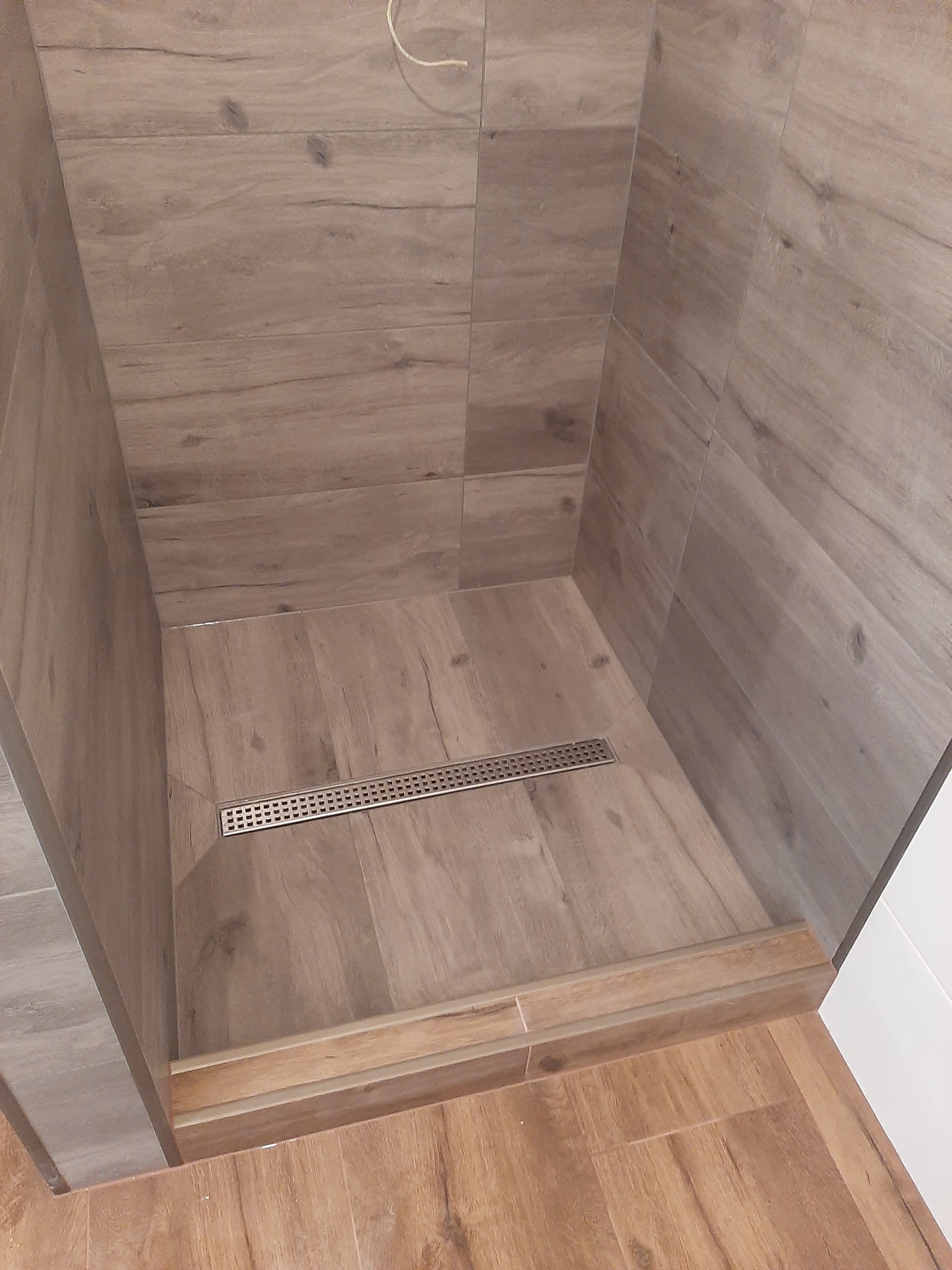 Wykończony brodzik prysznicowy z odpływem liniowym, wyłożony płytkami imitującymi drewno, z widocznym fragmentem podłogi łazienki.