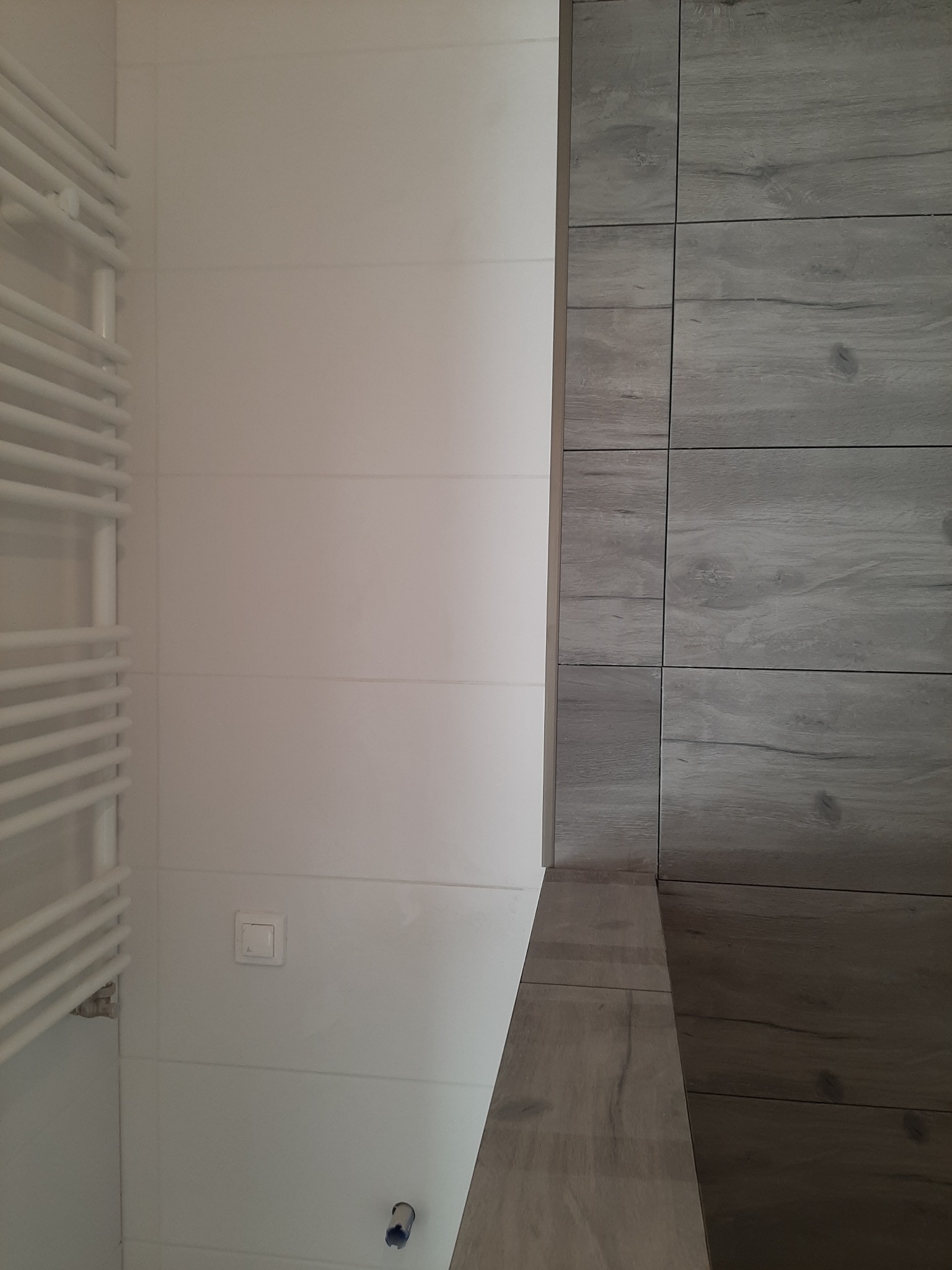 Wykończona ściana w łazience z białymi płytkami i grzejnikiem drabinkowym po lewej stronie oraz płytkami imitującymi drewno po prawej stronie. Widoczny biały włącznik światła i fragment instalacji...