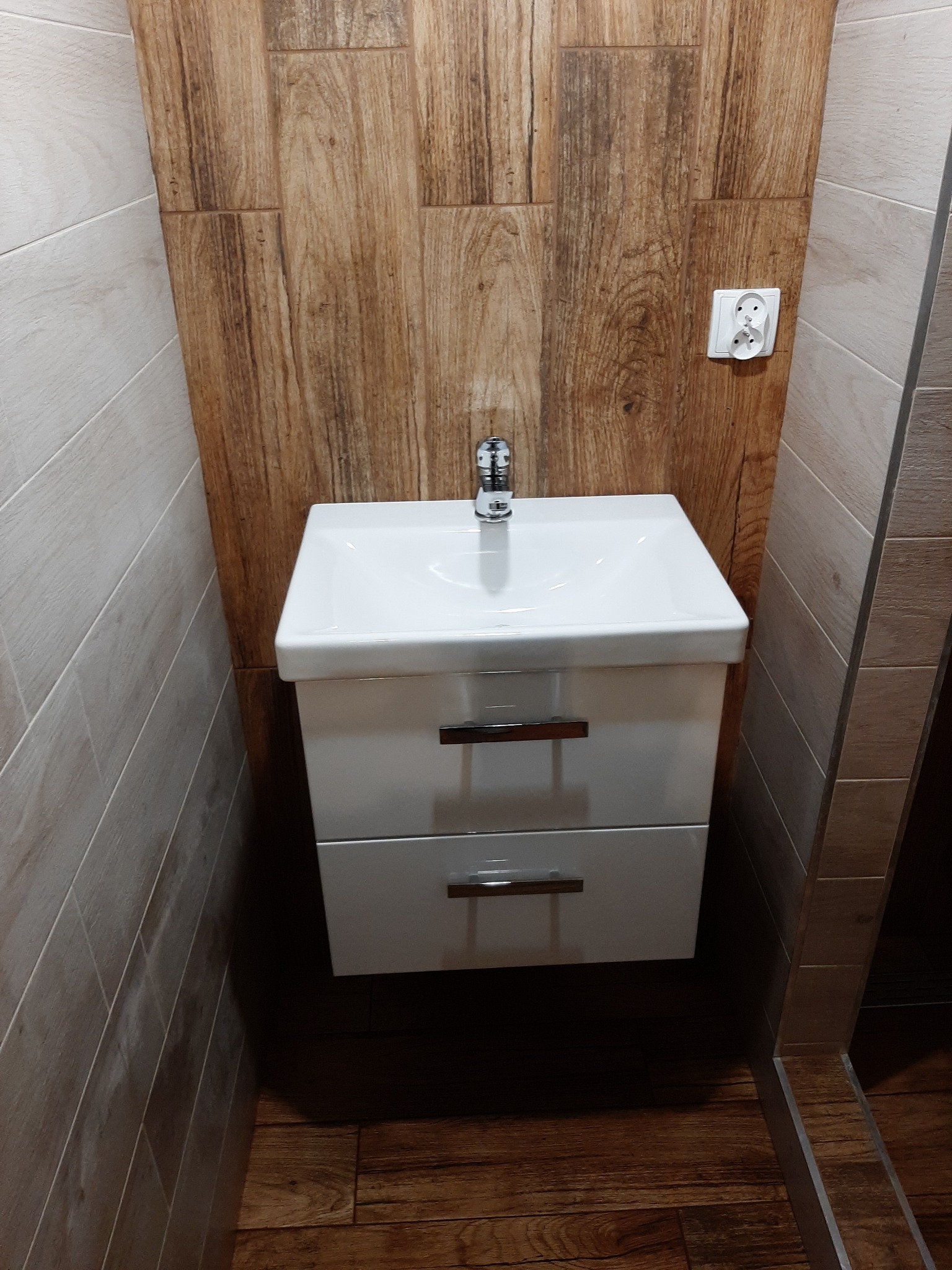 Nowoczesna łazienka z białą szafką pod umywalką z dwoma szufladami, chromowaną baterią, drewnopodobną okładziną ścienną i gniazdkiem elektrycznym.