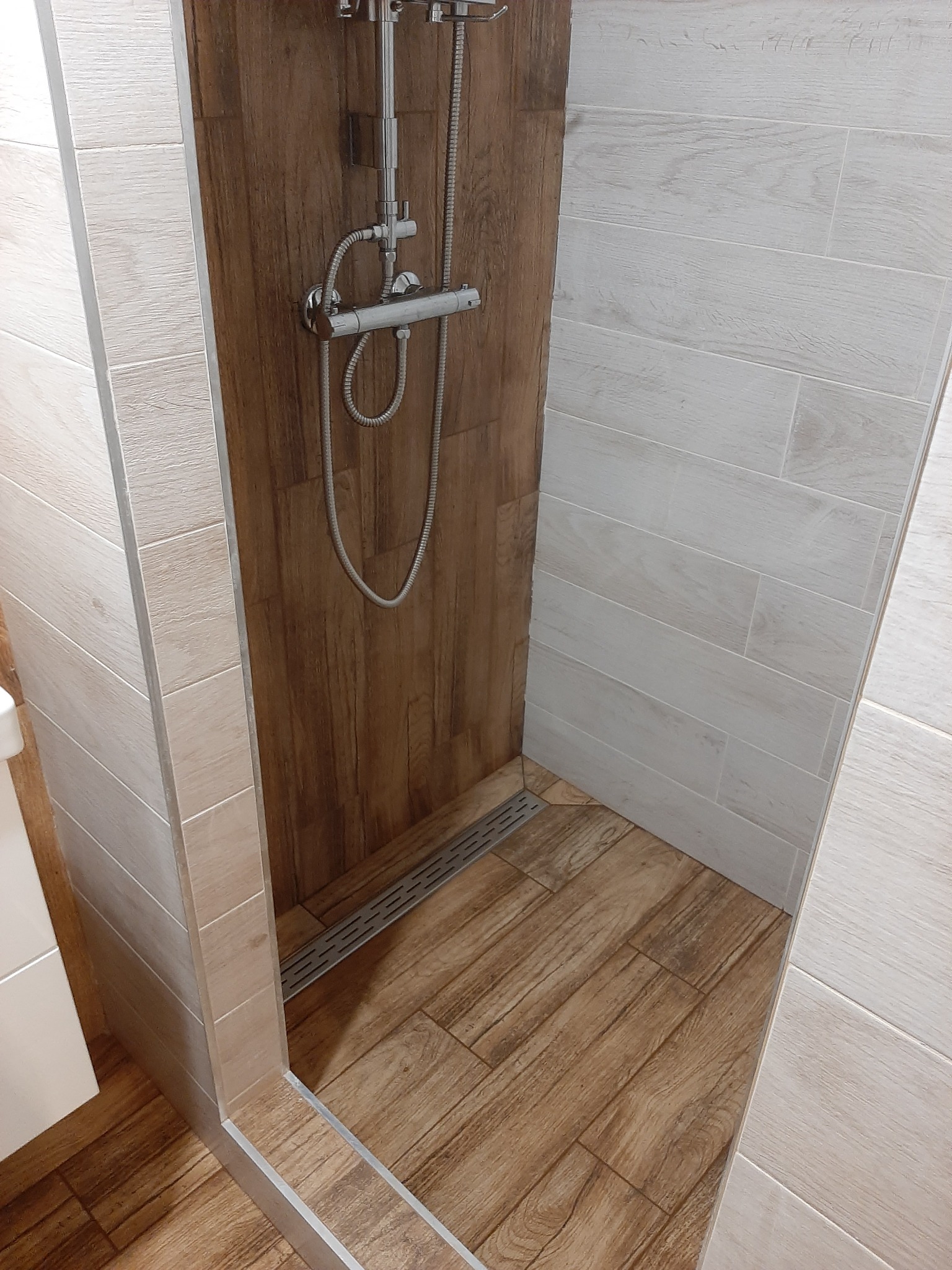 Nowoczesna kabina prysznicowa z odpływem liniowym, wykończona płytkami imitującymi drewno w dwóch odcieniach, jasne na ścianach i ciemniejsze na podłodze oraz tylnej ścianie kabiny.