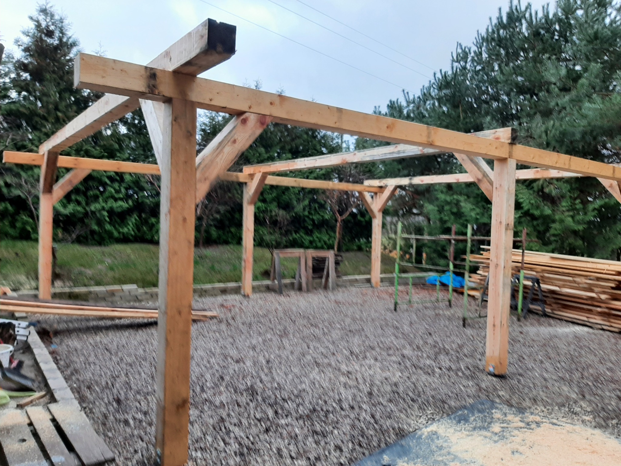 Drewniana konstrukcja altany w trakcie budowy, widoczne słupy i belki, w tle zielone drzewa, na ziemi rozsypane wióry.