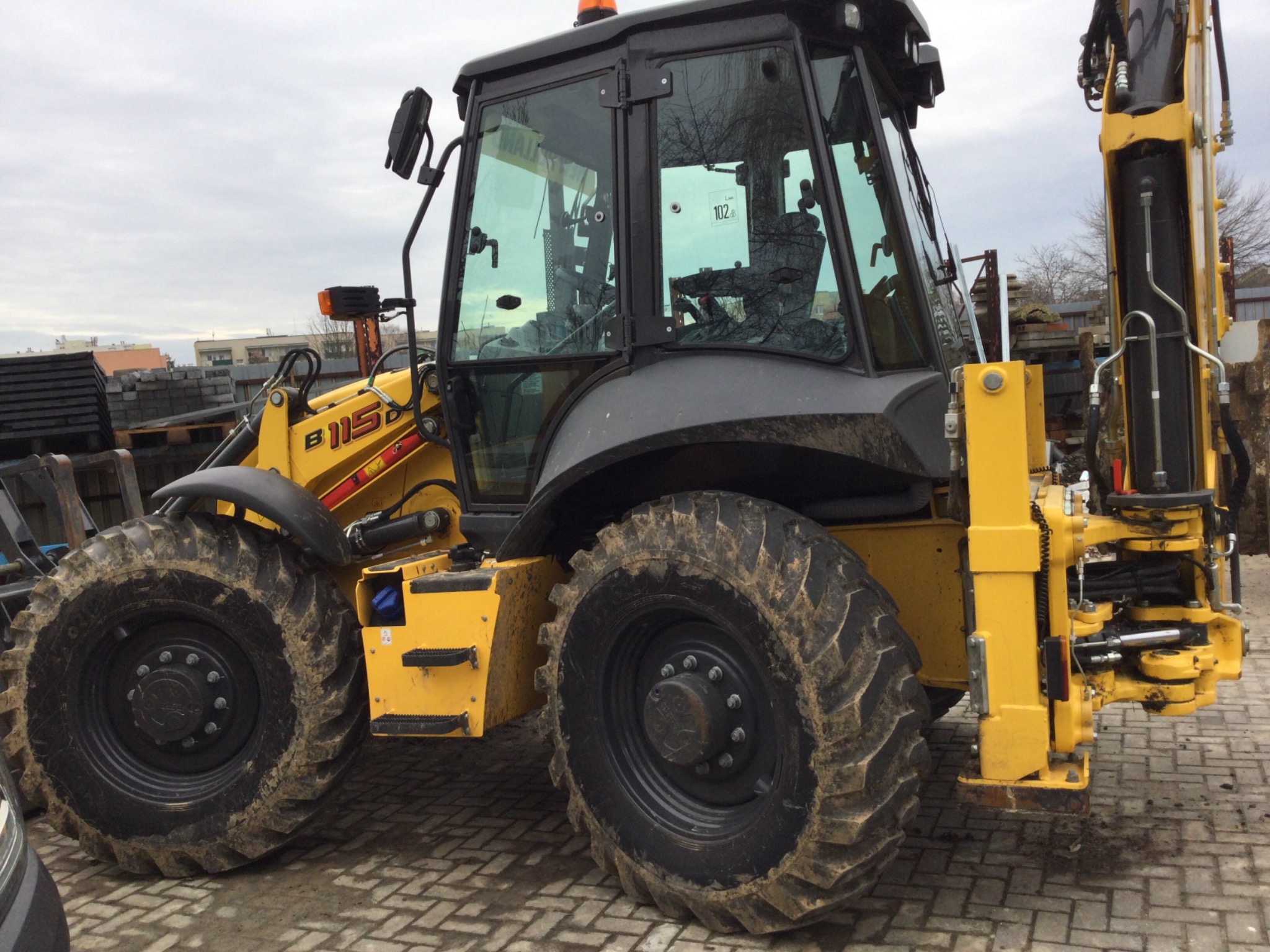 Żółto-szara koparko-ładowarka New Holland B115D z brudnymi oponami na kostce brukowej, z widocznymi elementami hydraulicznymi i naklejką '102' na szybie kabiny.