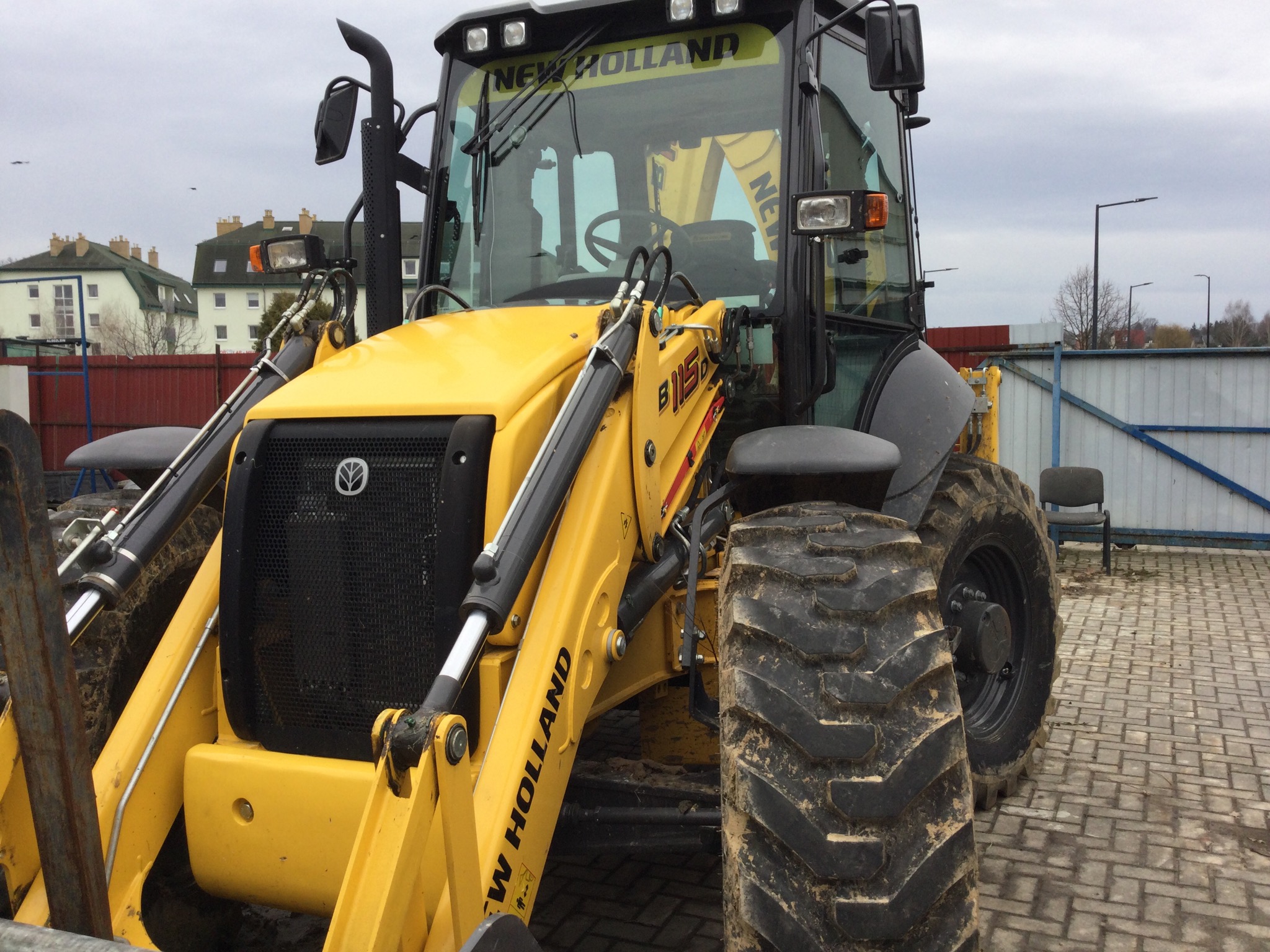 Żółta koparko-ładowarka New Holland B115B, widok z przodu, z elementami hydraulicznymi i oponami z wyraźnym bieżnikiem, na tle budynków i pochmurnego nieba.