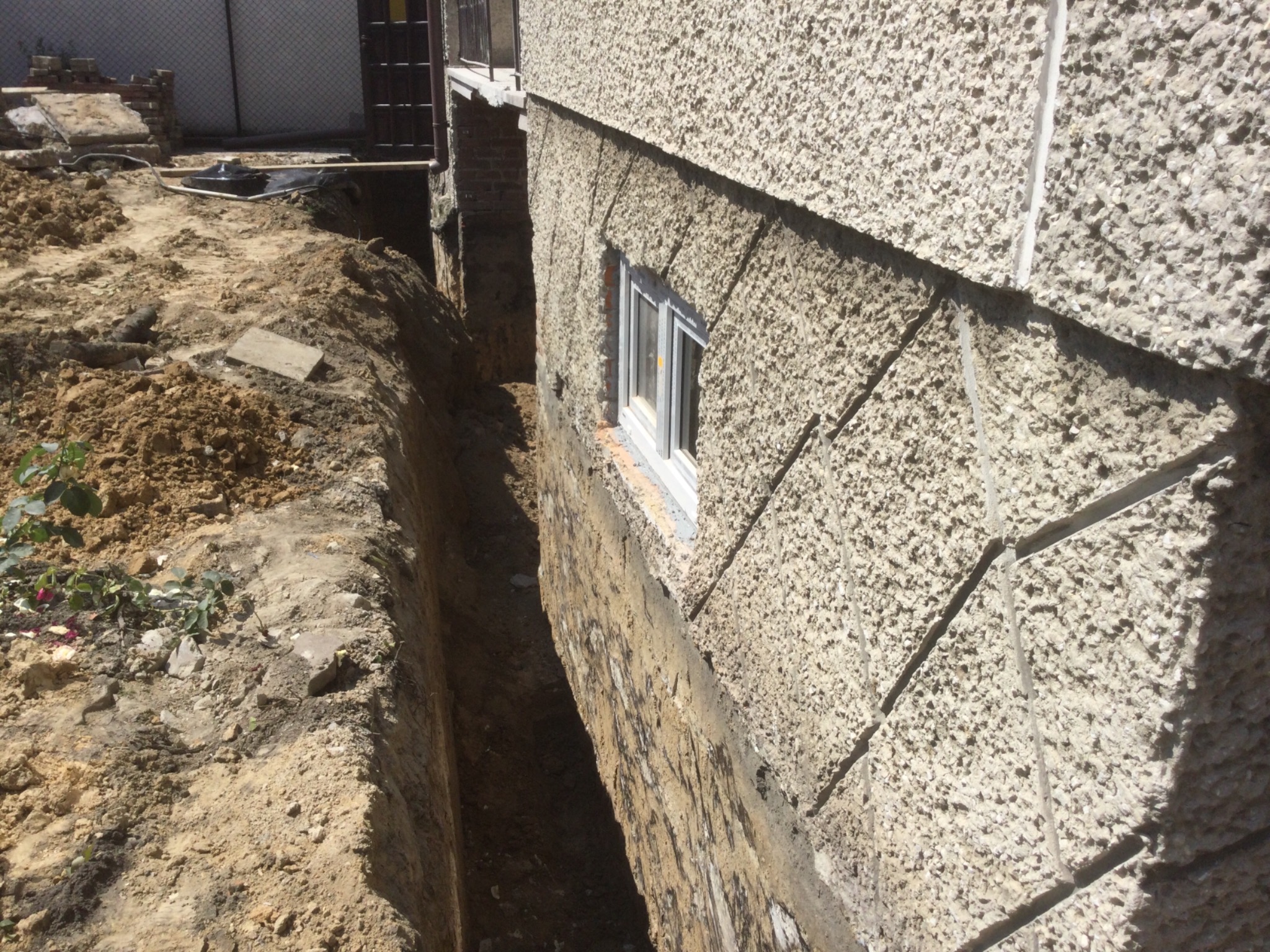 Wykop wzdłuż ściany budynku z oknem, odsłonięty fundament. Widoczne ślady wilgoci na fundamencie. Ziemia usypana po jednej stronie wykopu.
