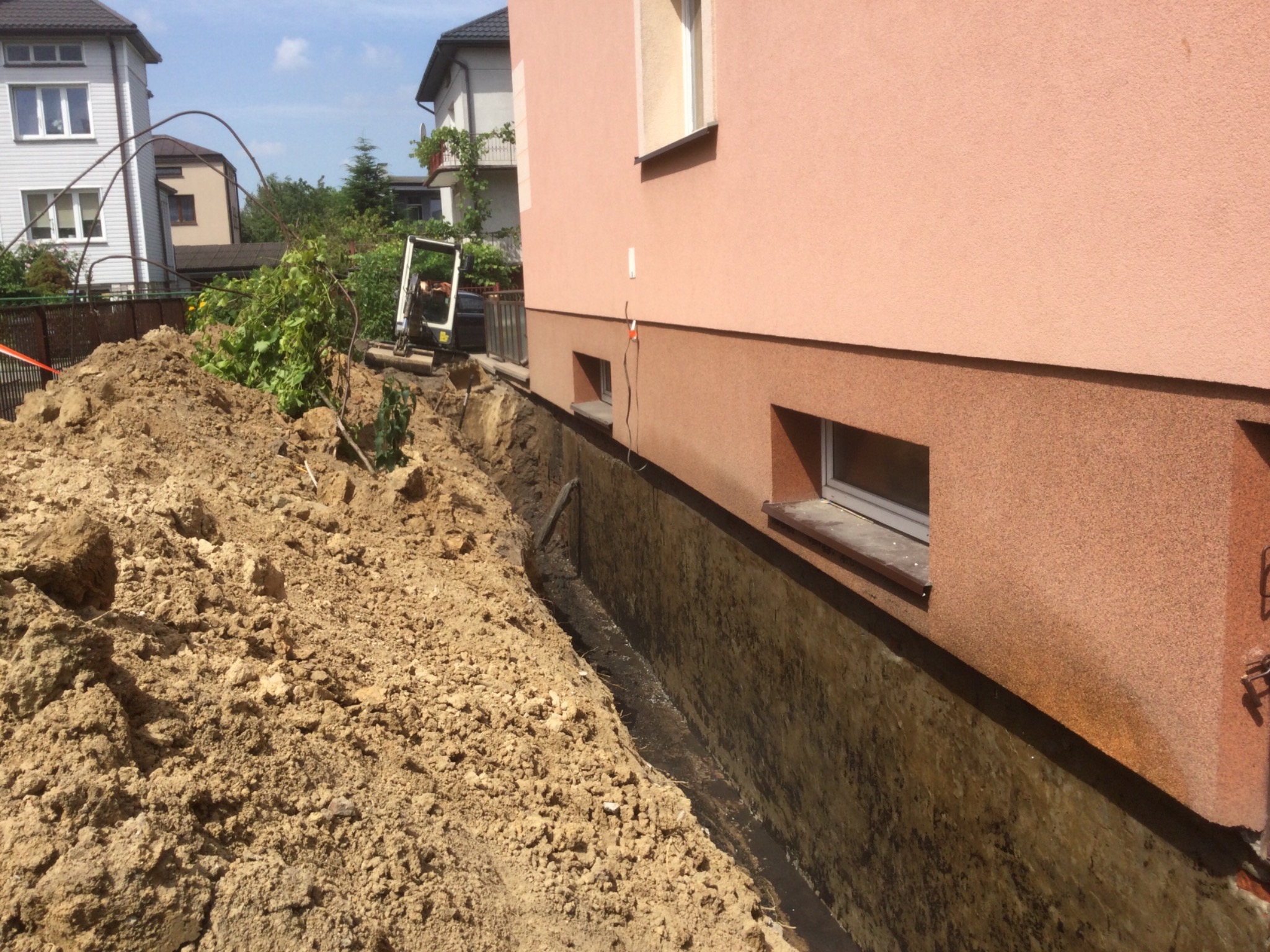 Wykop wzdłuż ściany budynku z widocznym fragmentem minikoparki w tle.